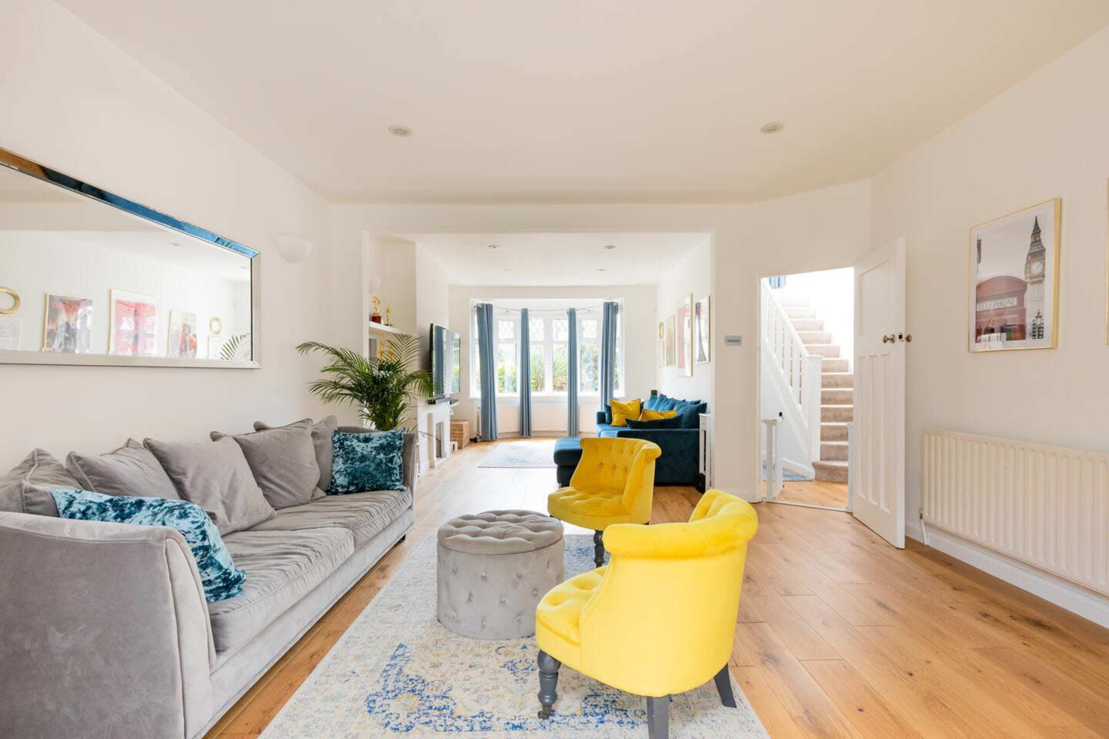 A modern living room features a gray sofa, yellow armchairs, a round ottoman, and large windows overlooking a garden.