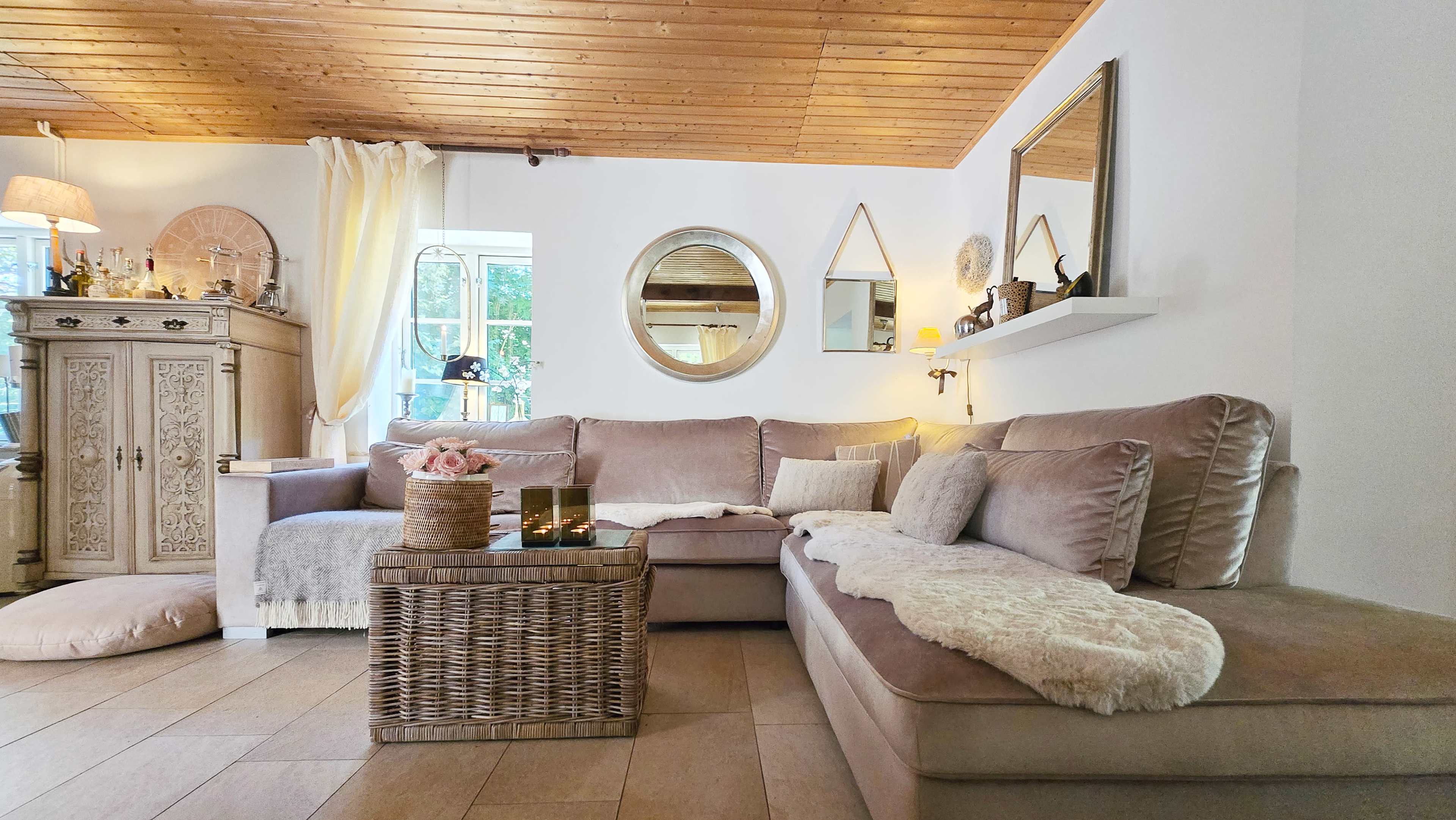 The image shows a cozy living room featuring a sectional sofa with a light-colored throw, a wicker coffee table, and decorative elements on the walls, including mirrors and shelves.