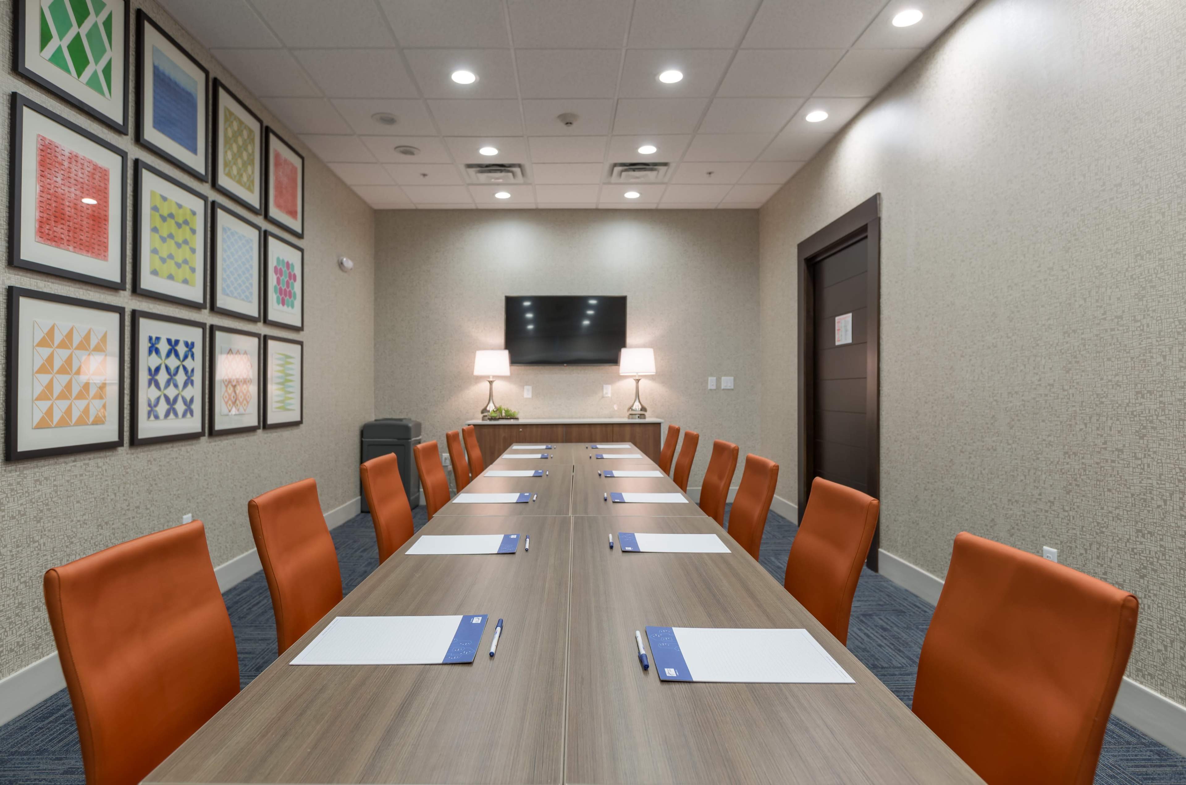 A conference room features a long wooden table with orange chairs, framed artwork on the walls, and a television mounted at the far end.