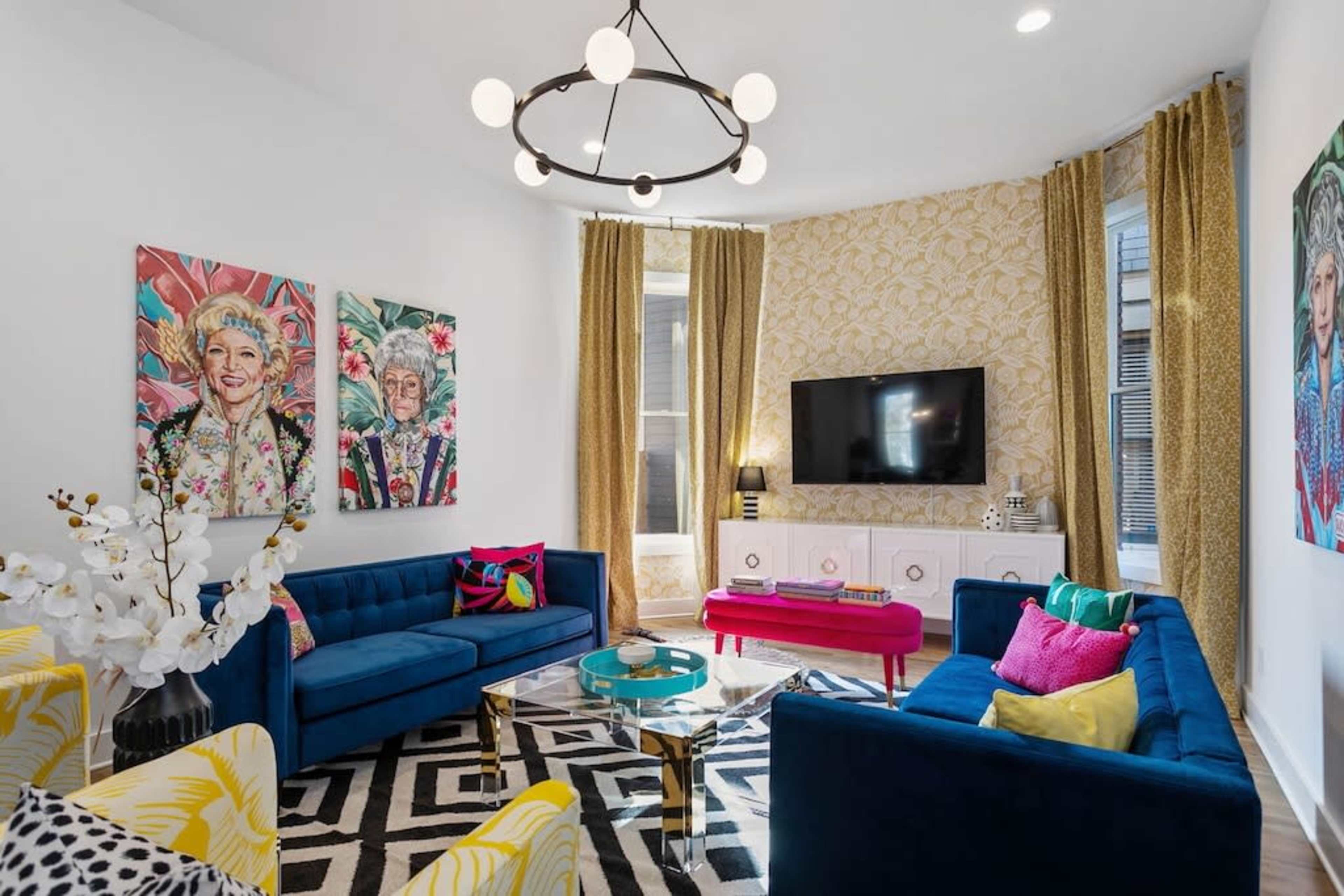 The image shows a brightly decorated living room featuring blue sofas, colorful cushions, patterned curtains, and wall art, with a television mounted above a white cabinet.