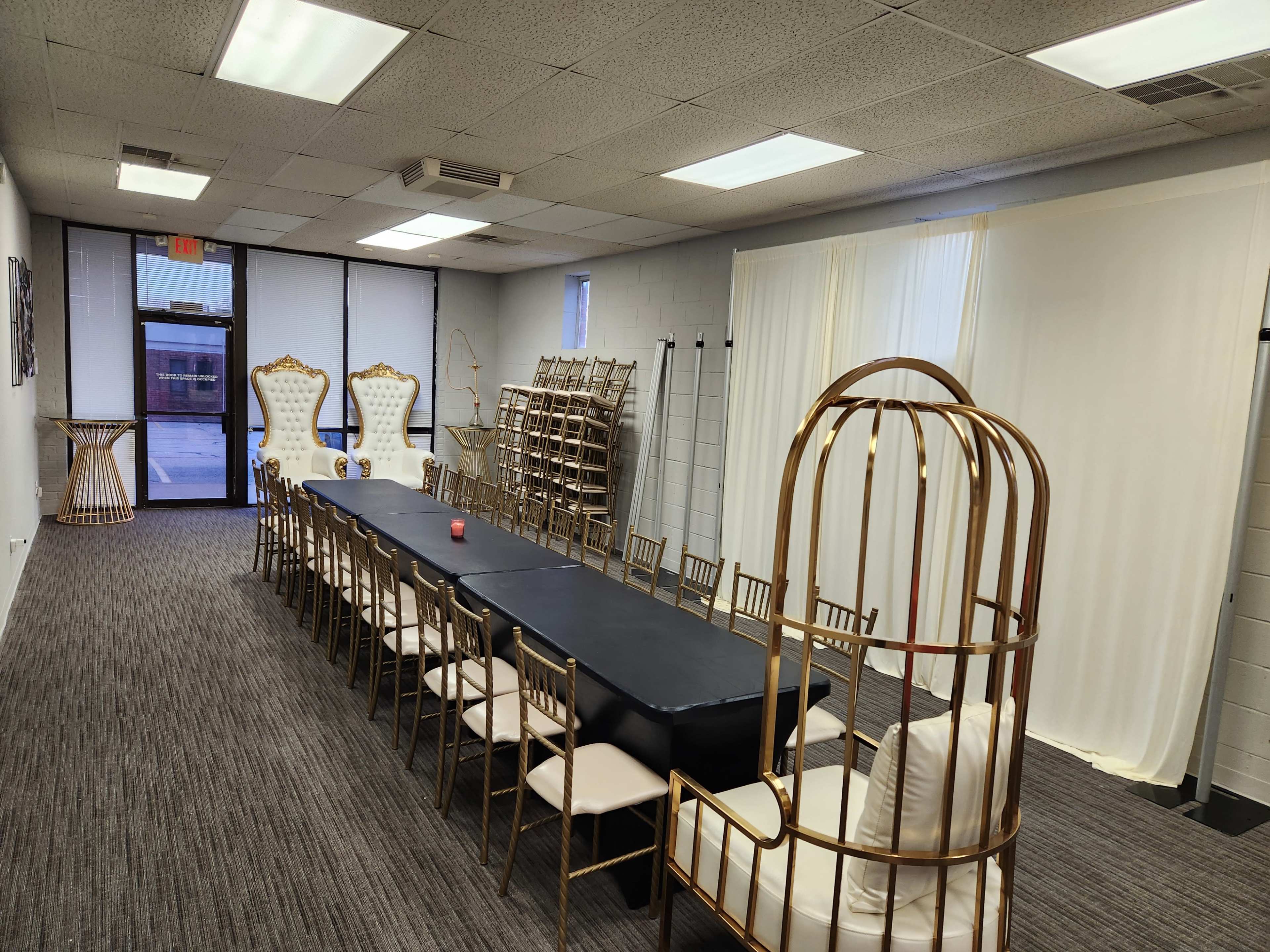 The room features a long black table with chairs arranged around it, flanked by decorative gold chairs and several standing racks in a modern setting.