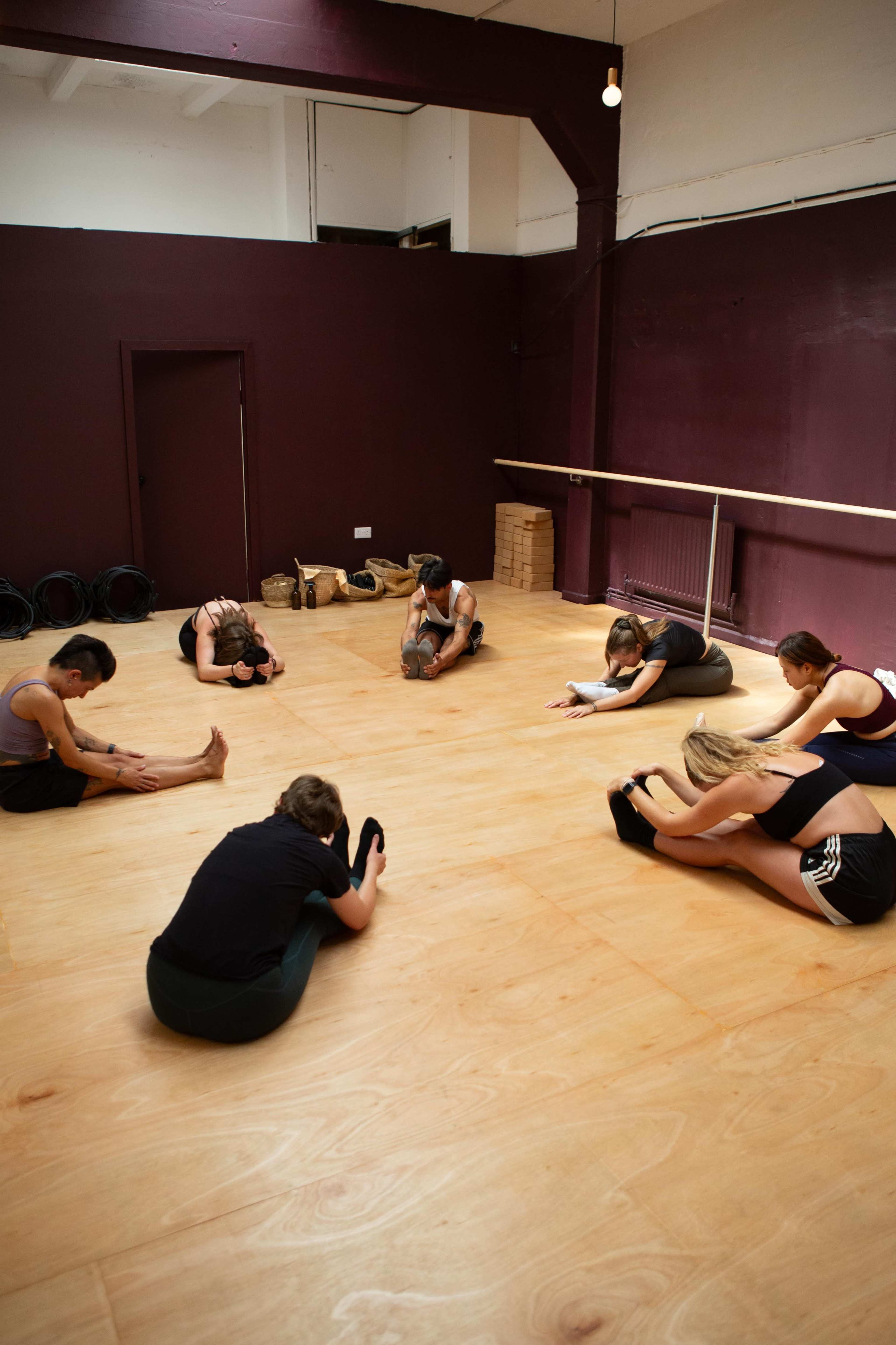 A group of six individuals are performing stretching exercises on a wooden floor in a spacious studio with maroon walls.