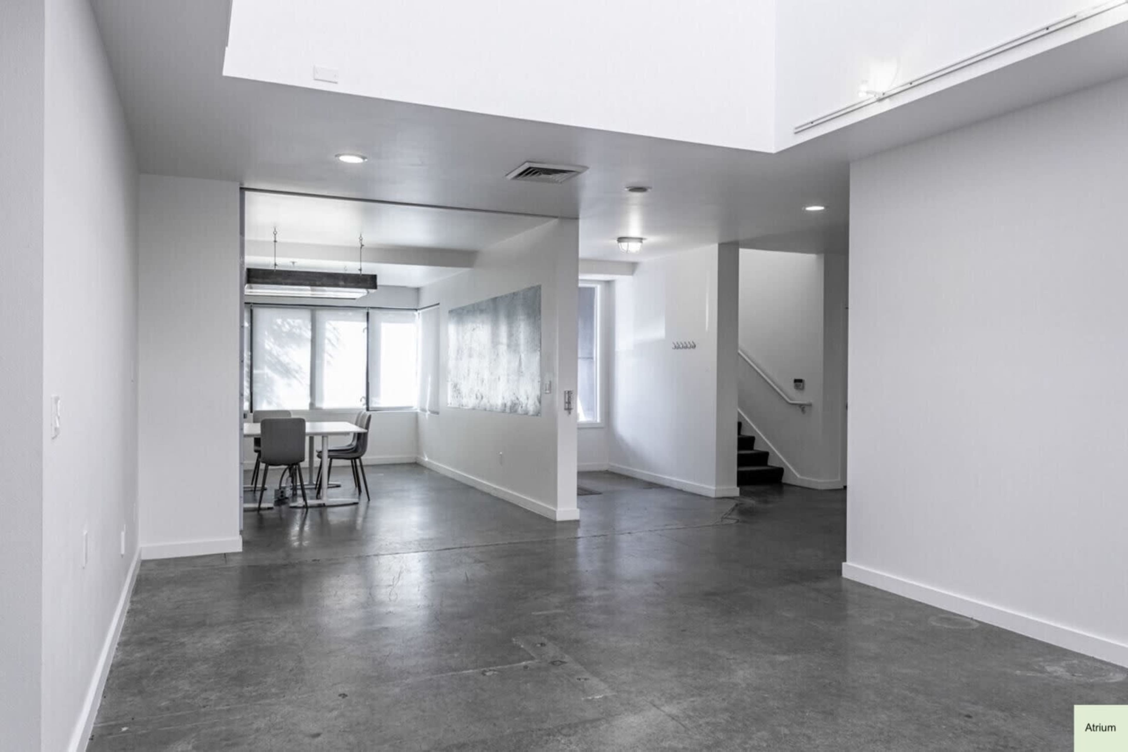 The image shows an interior space featuring a concrete floor, a dining area with a table and chairs, a large wall possibly used for presentation, and a staircase leading to another level.