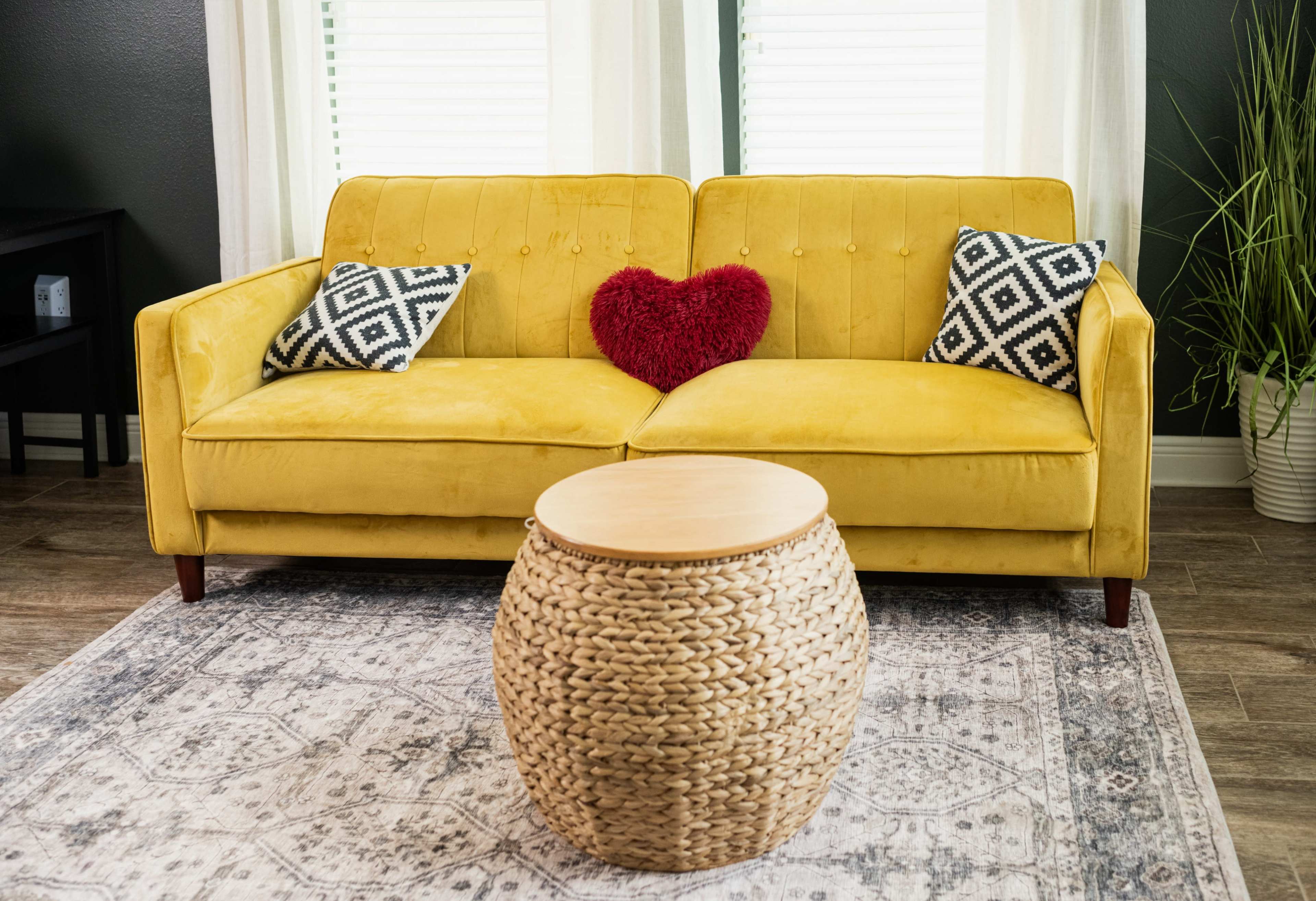 A yellow couch with black and white patterned pillows sits on a patterned rug, next to a round woven coffee table, in a well-lit room with window curtains.