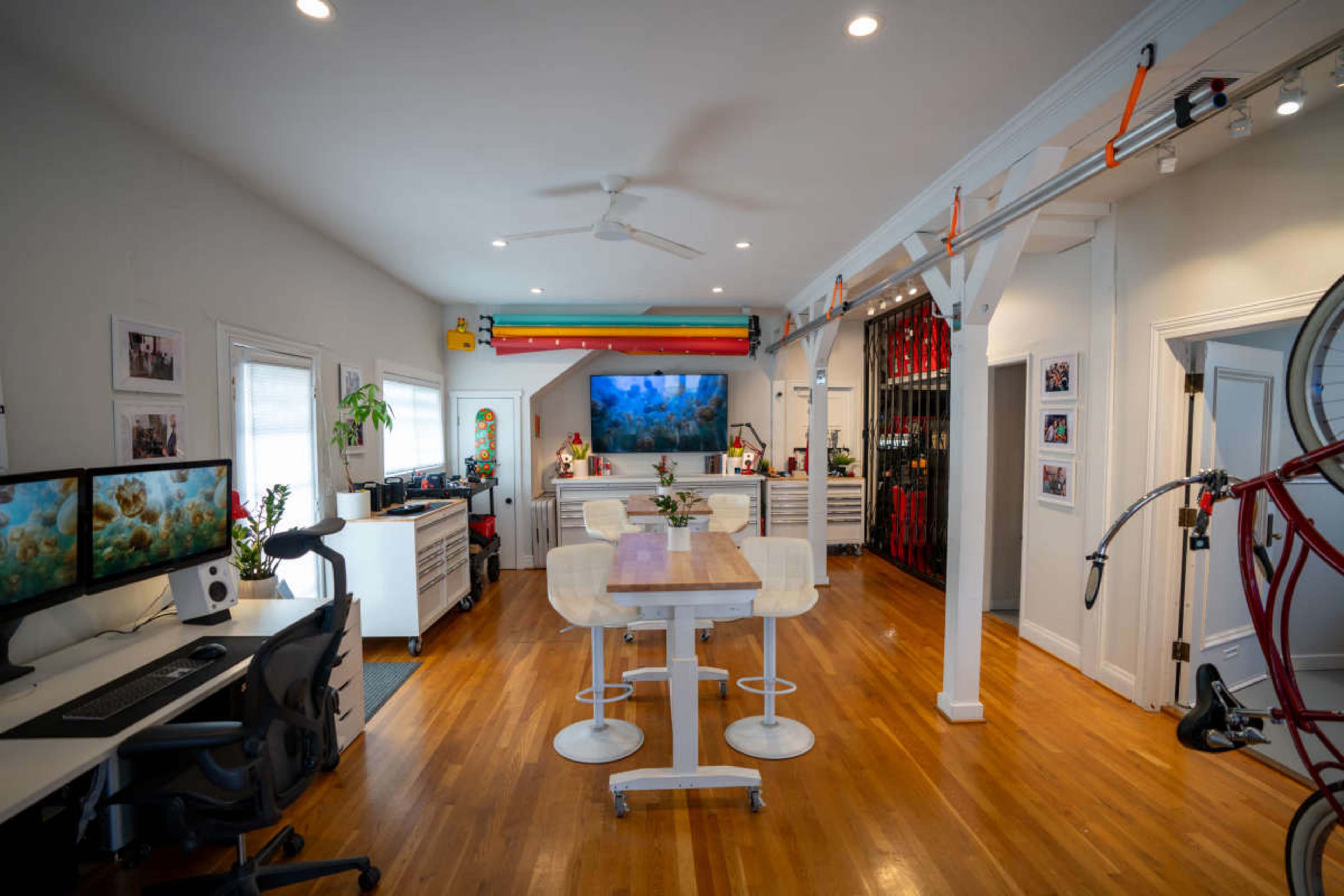 The image shows a modern open-concept office space featuring a wooden table, workstations with computers, and a large screen displaying an underwater scene, all surrounded by white walls and wooden flooring.