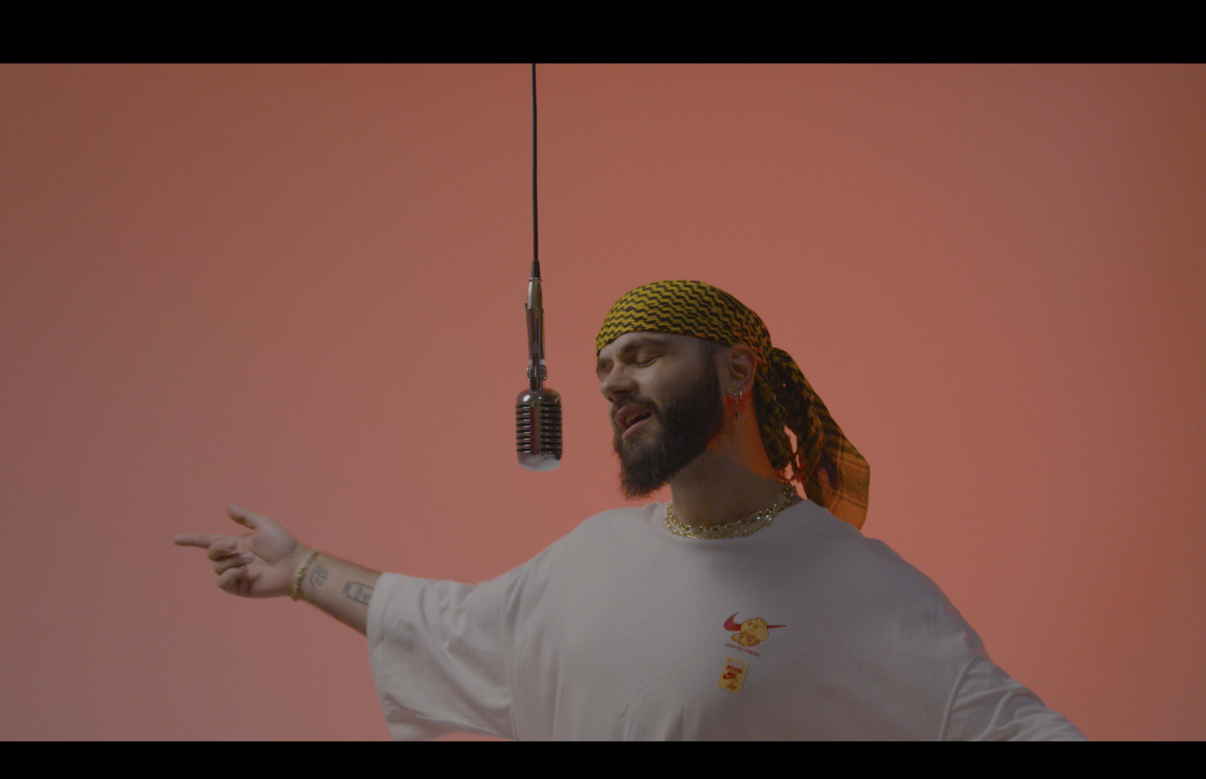 A man wearing a yellow bandana and a white oversized t-shirt sings into a vintage microphone against a gradient orange background.