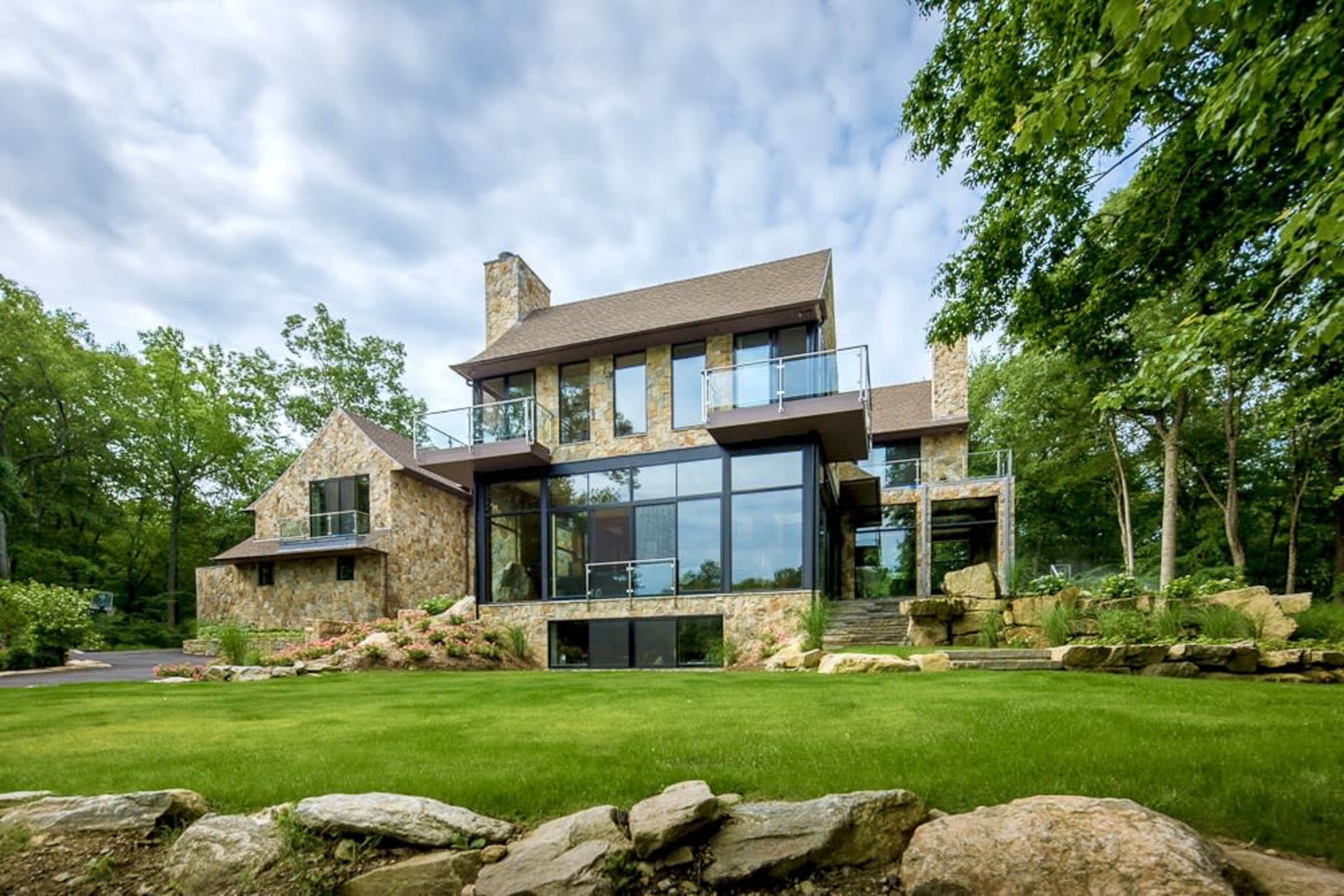 A modern stone and glass house is set against a backdrop of trees and a well-manicured lawn.