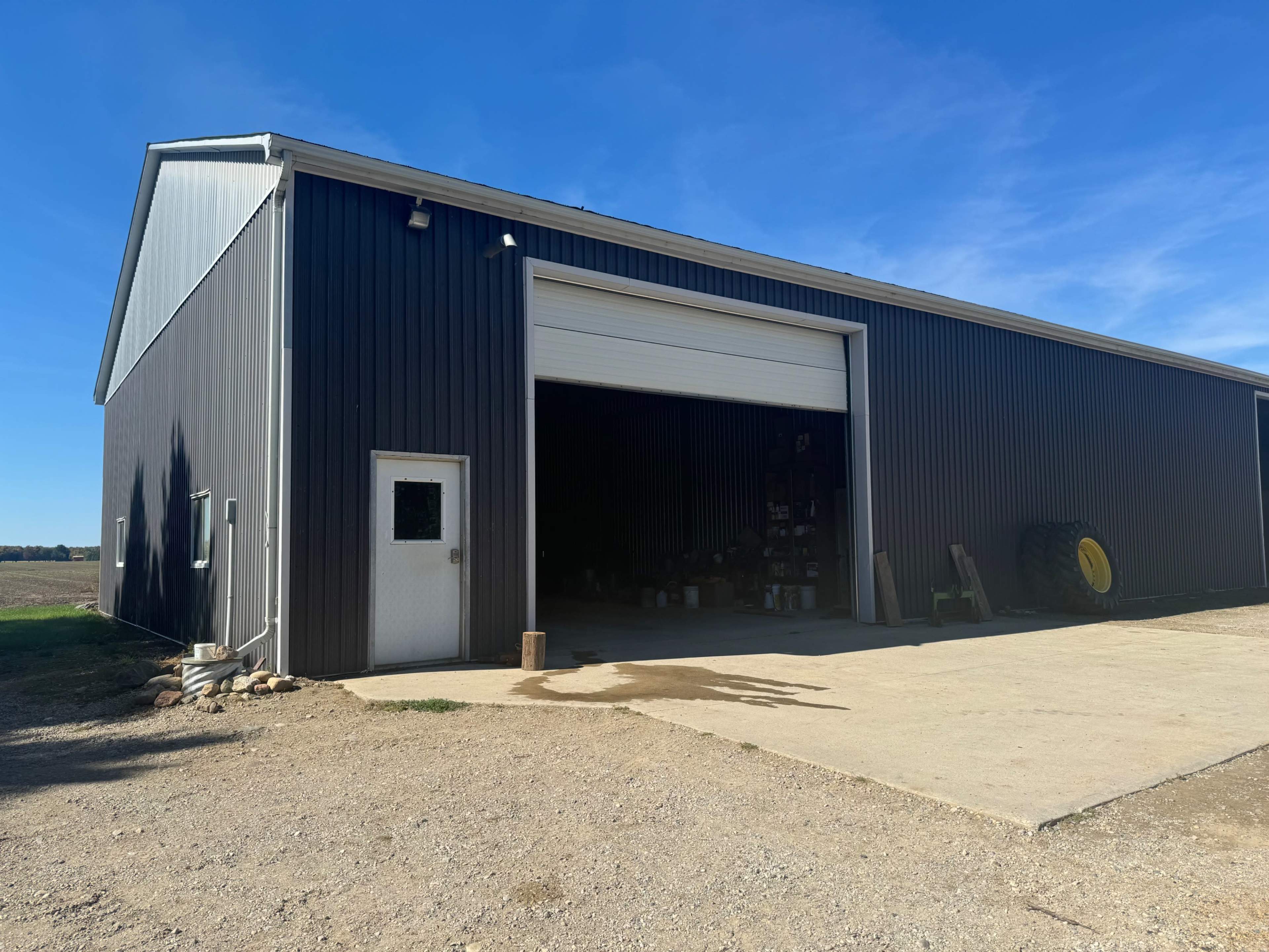 The image shows a large metal barn with a roll-up door, located on a gravel surface under a clear blue sky.