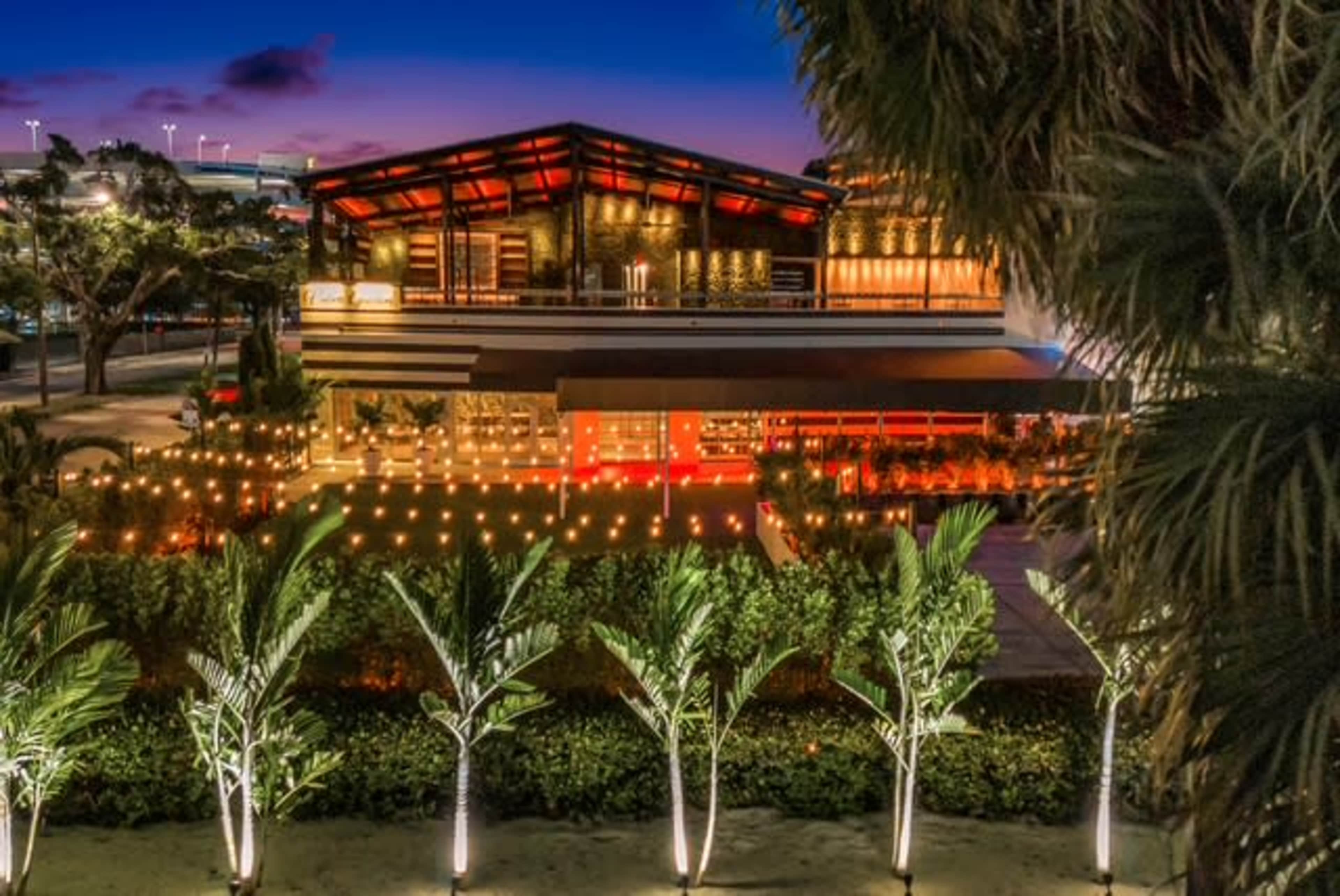 A modern building with a wooden exterior and illuminated landscaping sits amidst palm trees at dusk.