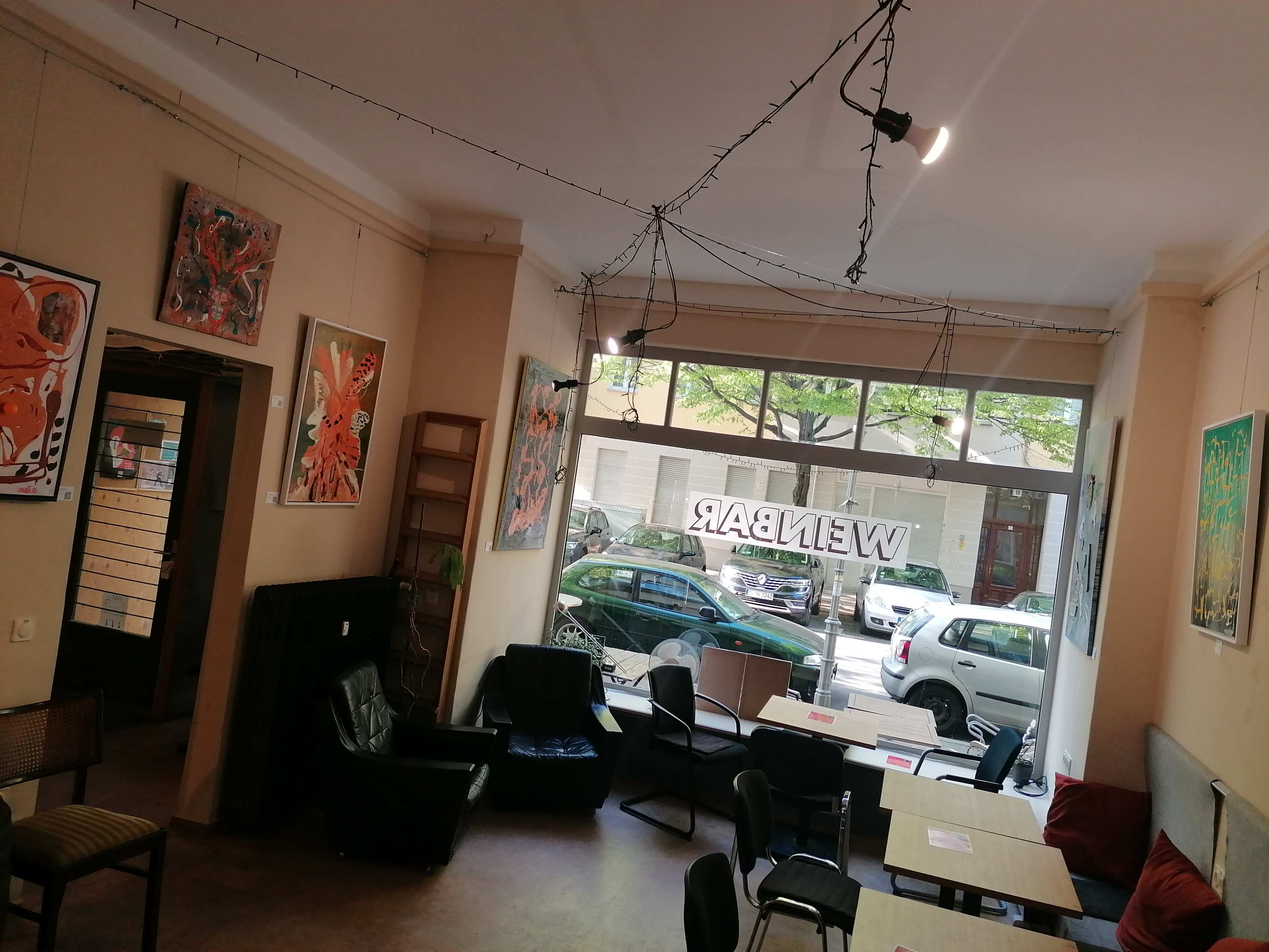 The image shows a café interior with art on the walls, tables and chairs set up for customers, and a large window facing the street outside.