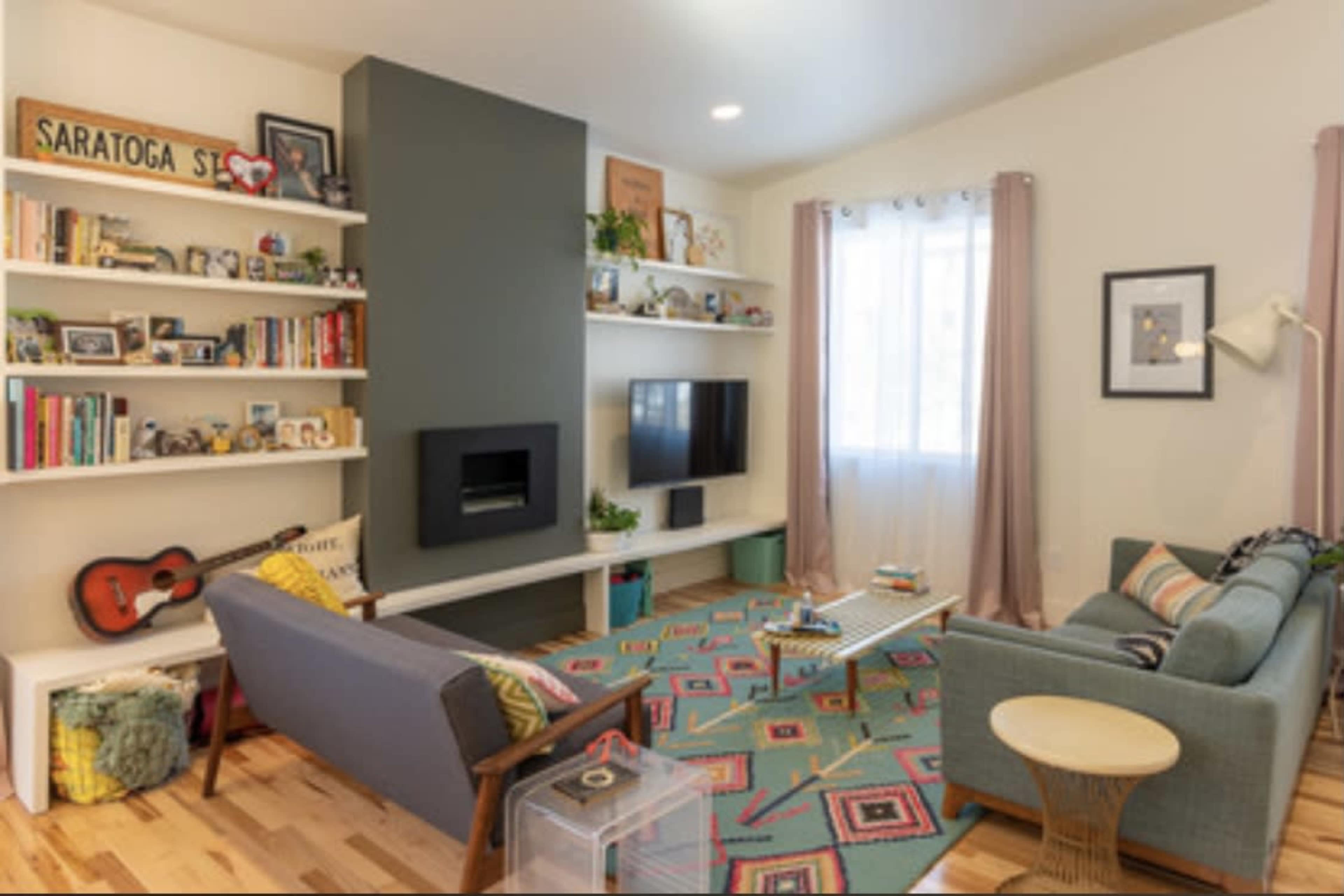 The living room features a grey accent wall, a modern fireplace, bookshelves filled with books and decor, a television mounted on the wall, and furniture arranged around a colorful area rug.
