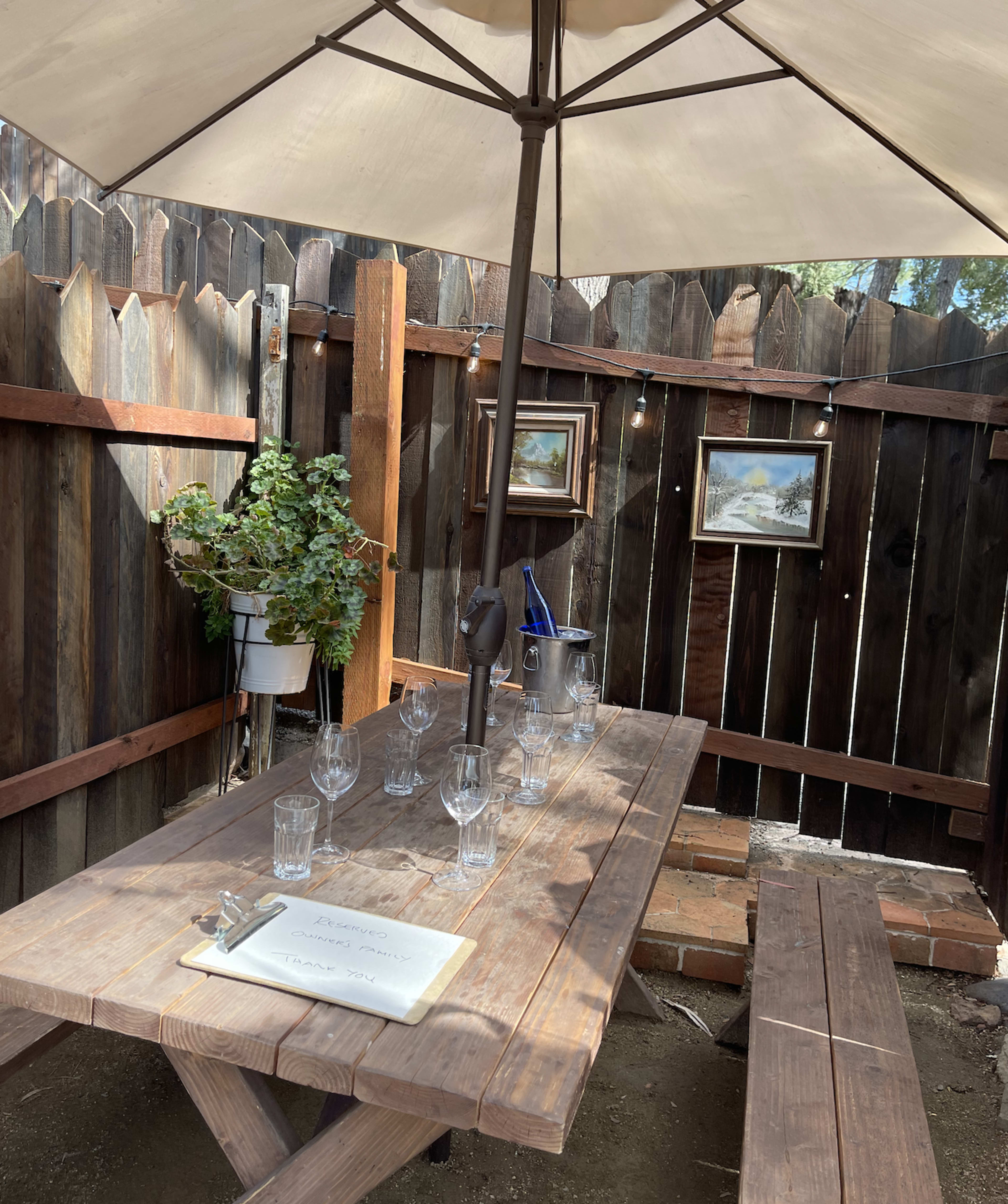 A wooden table is set under a patio umbrella, surrounded by a fenced area with hanging plants and framed pictures on the walls.