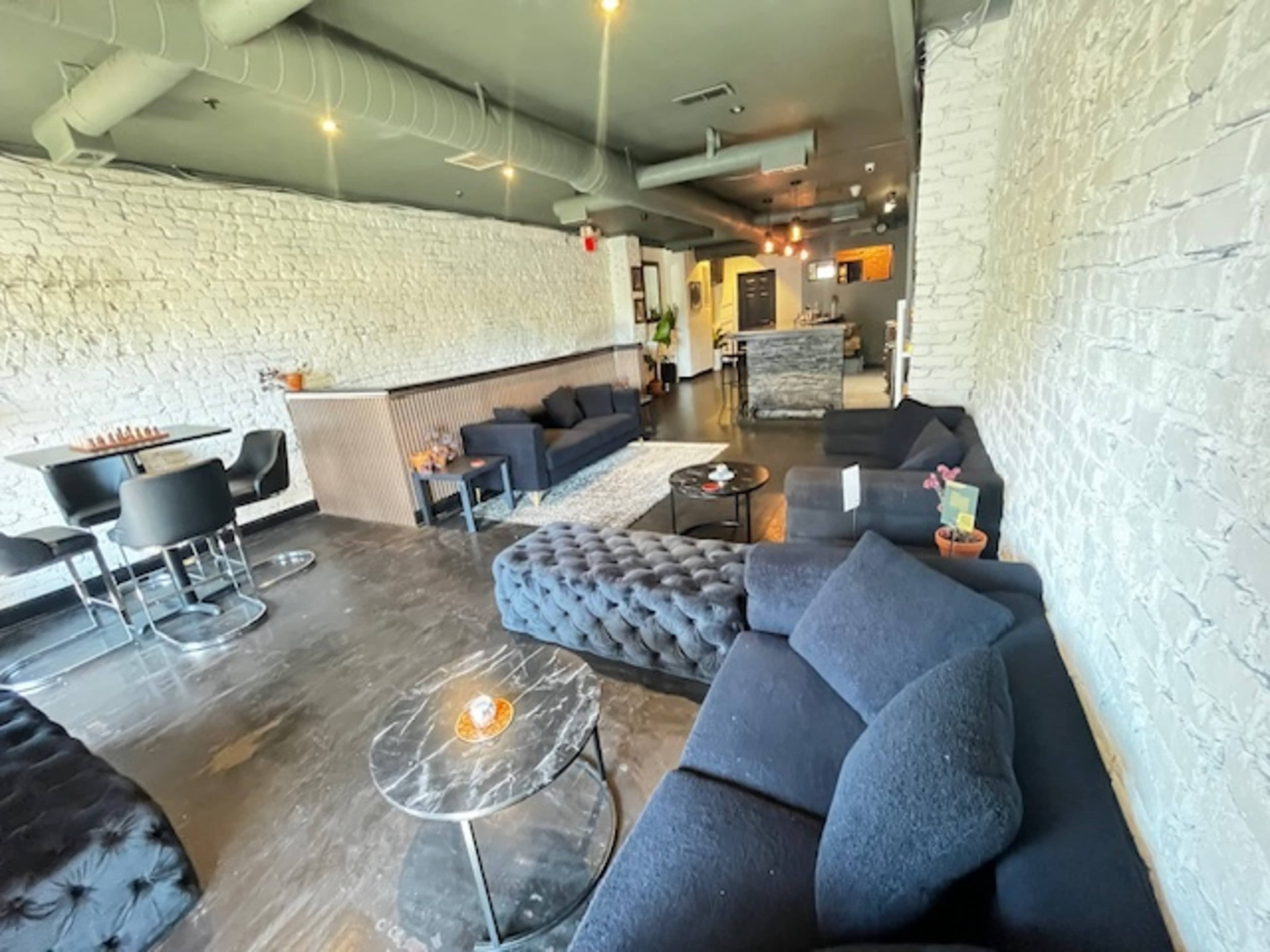 The image shows a modern lounge area featuring dark upholstered couches, a marble coffee table, and a bright, airy atmosphere with white brick walls.