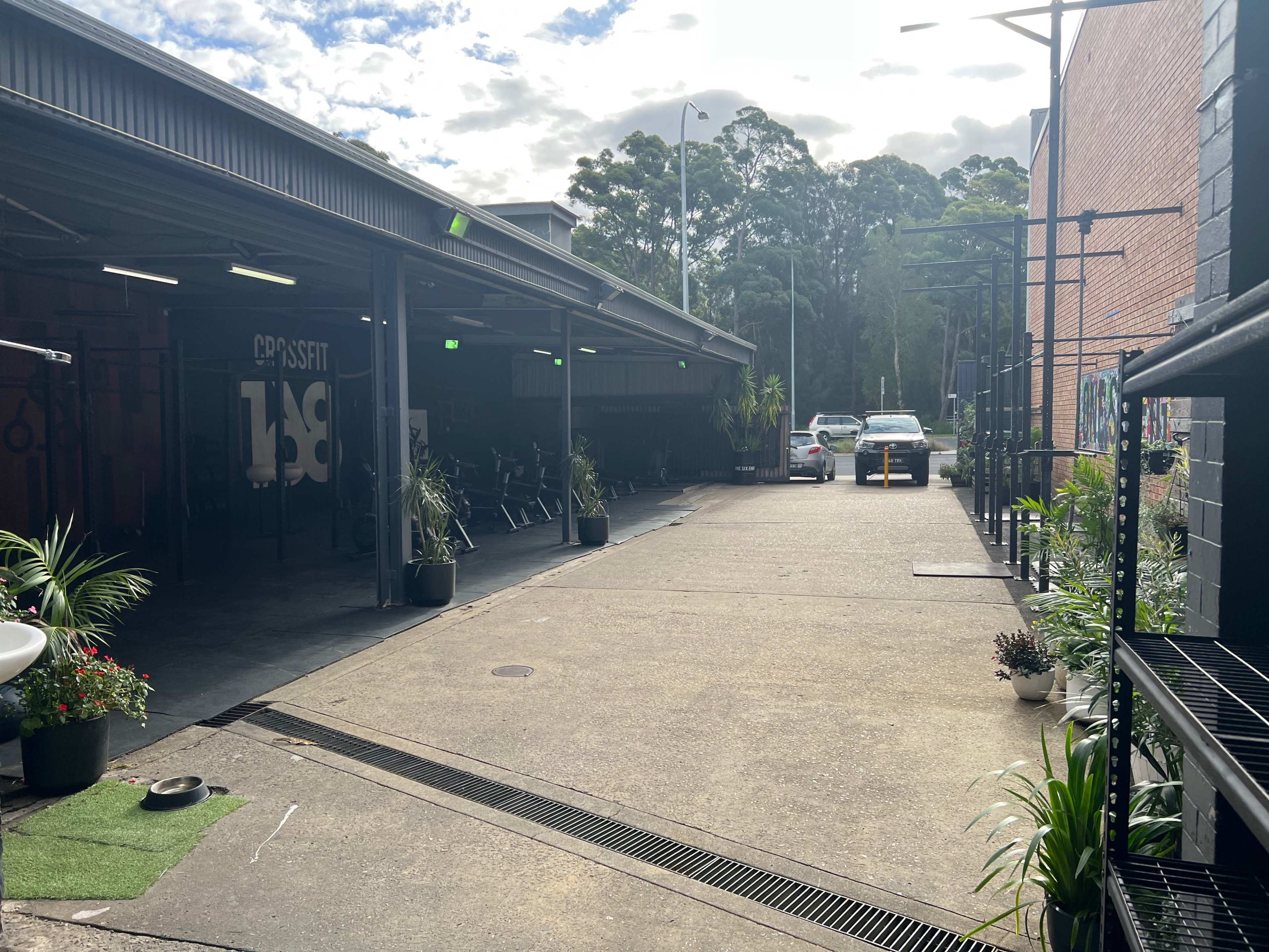 A wide, paved outdoor area flanked by buildings, with parked bicycles on one side and a few cars on the other, surrounded by plants and greenery.