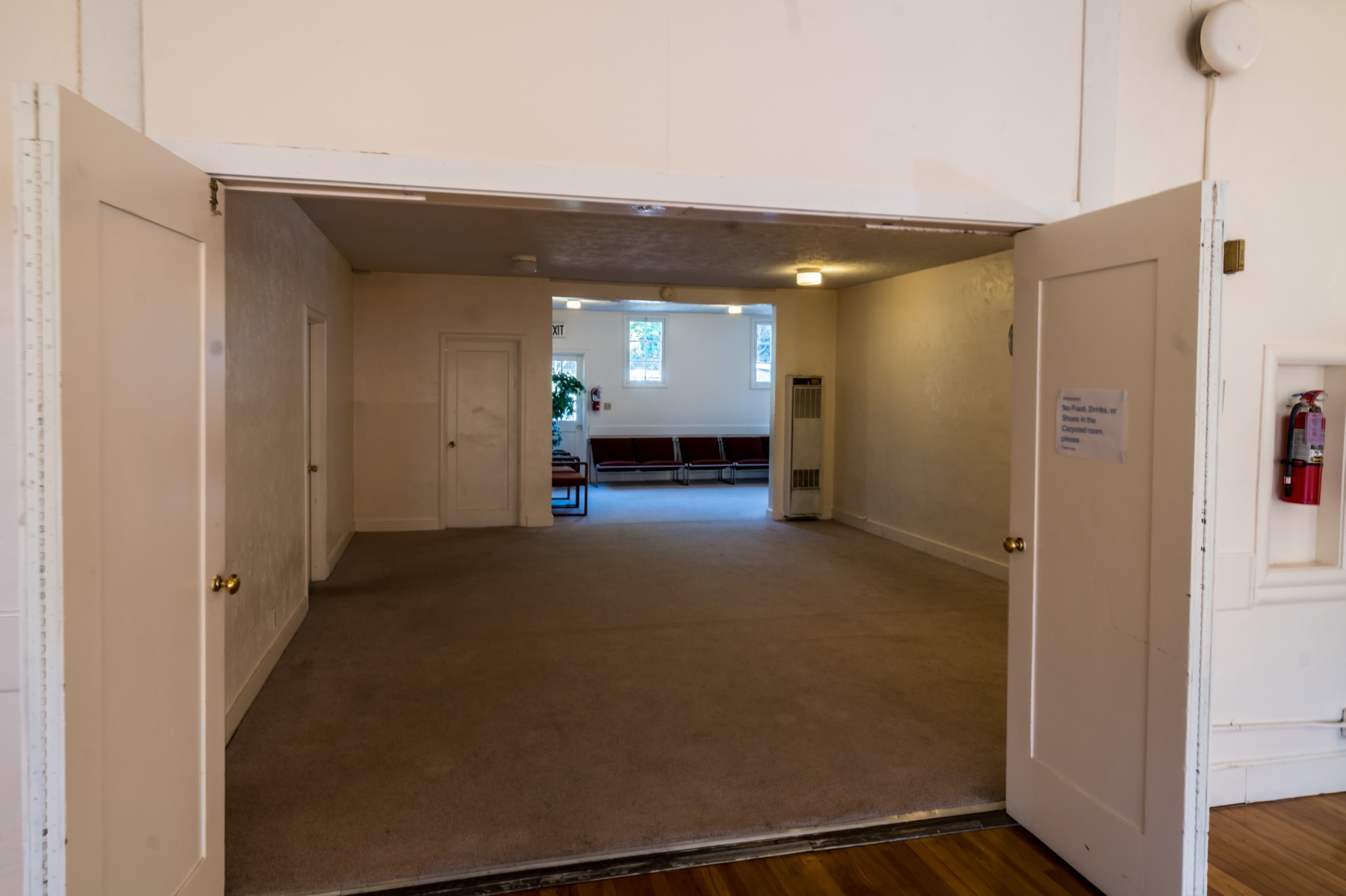 An empty, light-colored room with open double doors leading to another space featuring red chairs and a window.