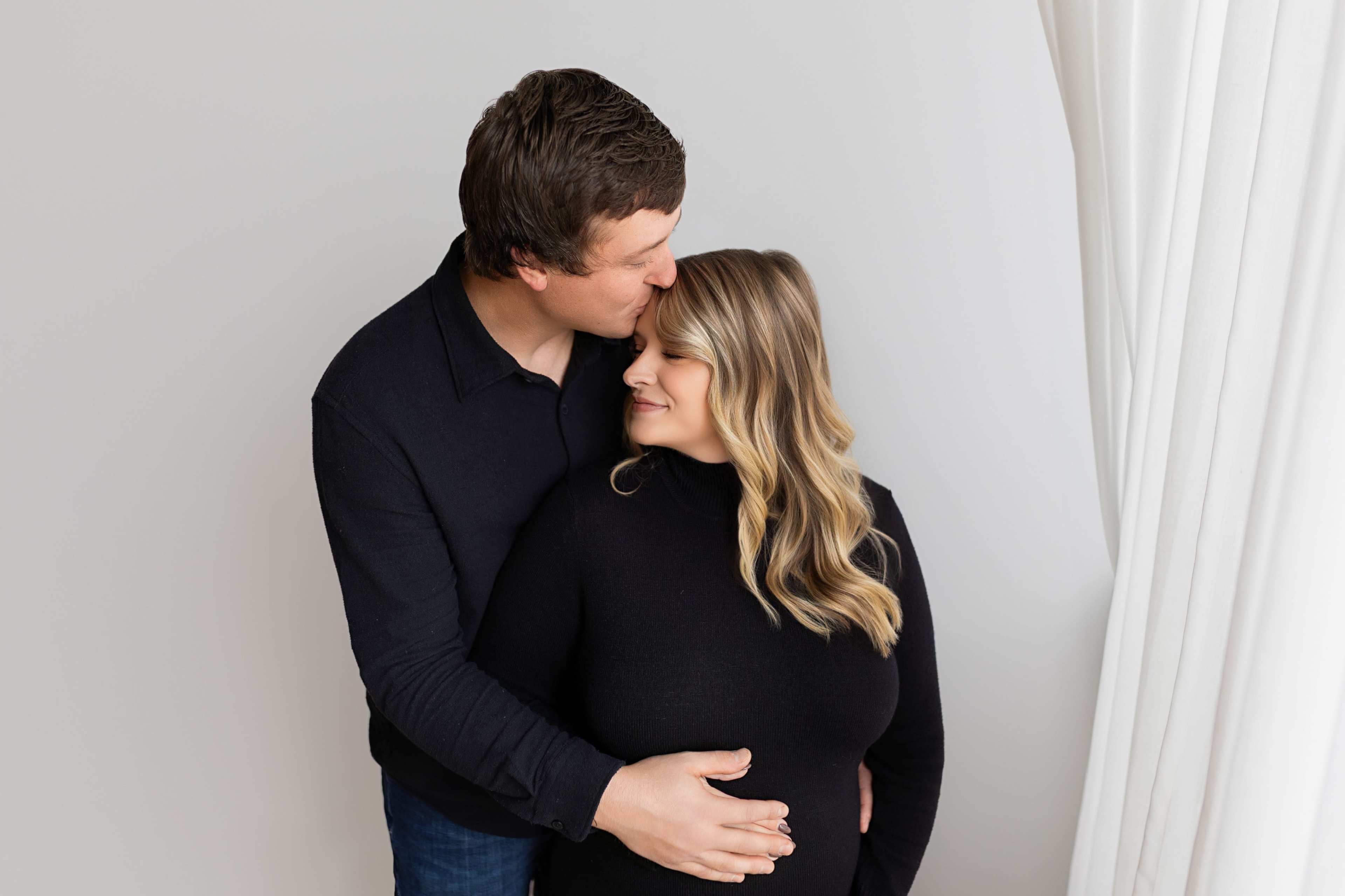 A couple embraces in front of a light-colored backdrop, with the man kissing the woman's forehead as she smiles gently.