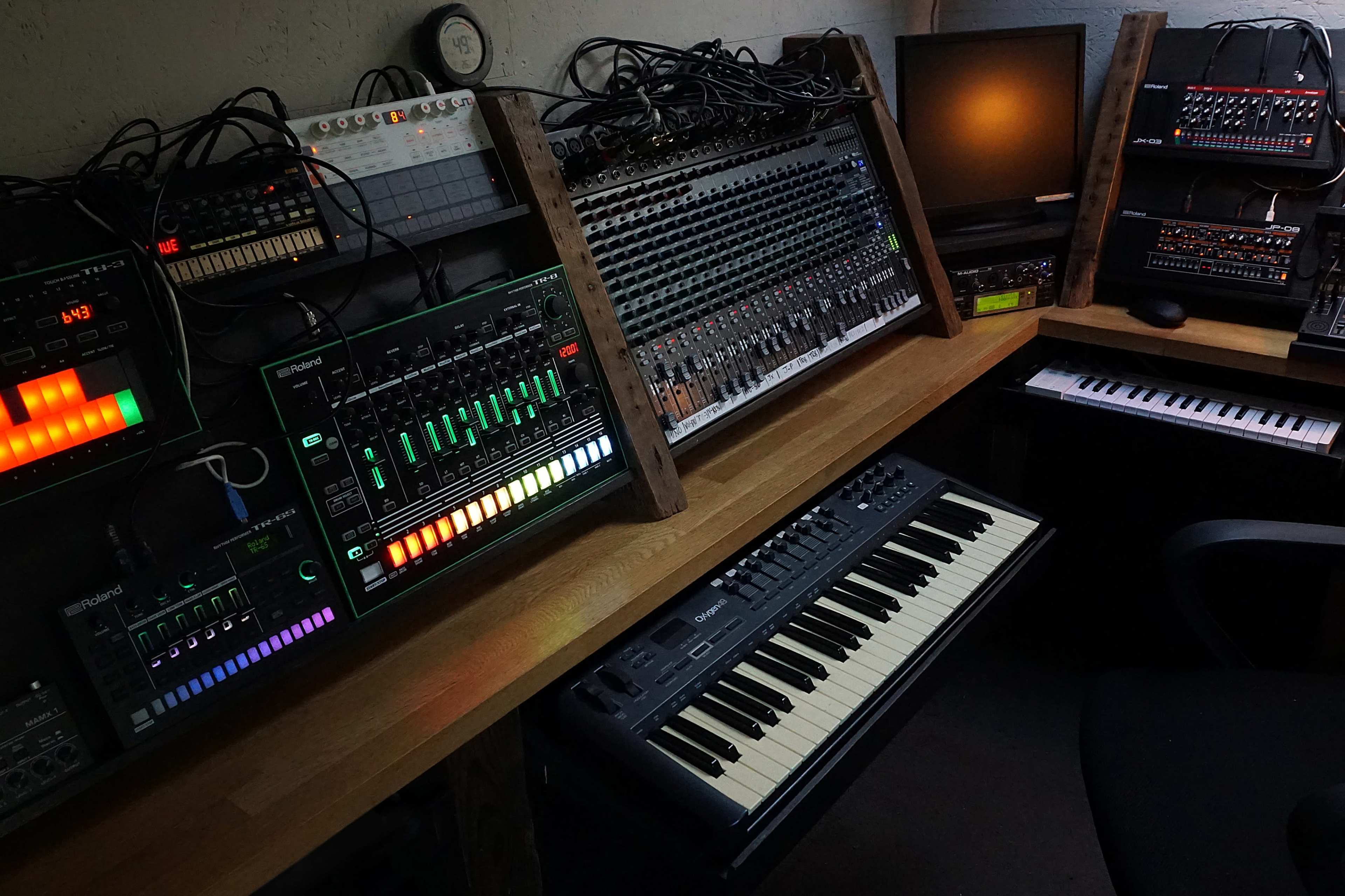 An arrangement of electronic musical instruments and equipment is set up on a wooden desk in a dimly lit room.