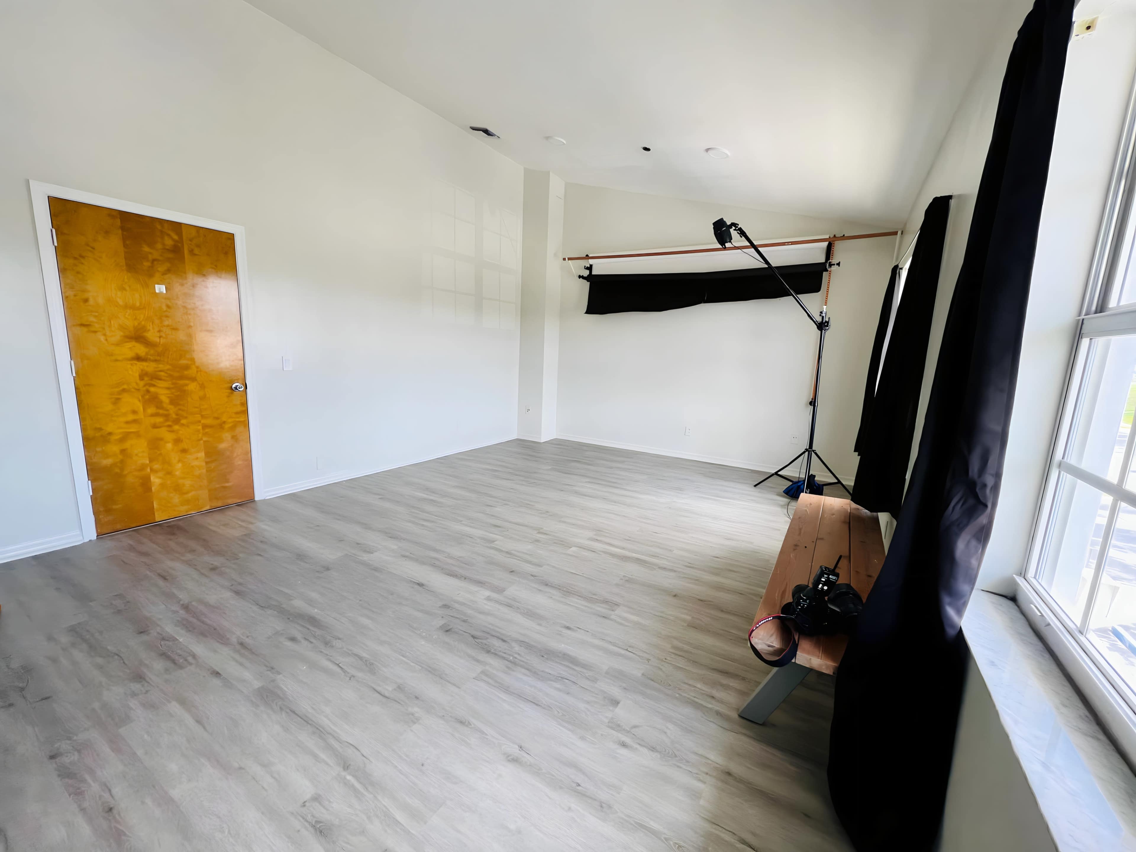 A spacious, empty studio with wooden flooring, a bench, and a backdrop setup along with a camera on the bench.