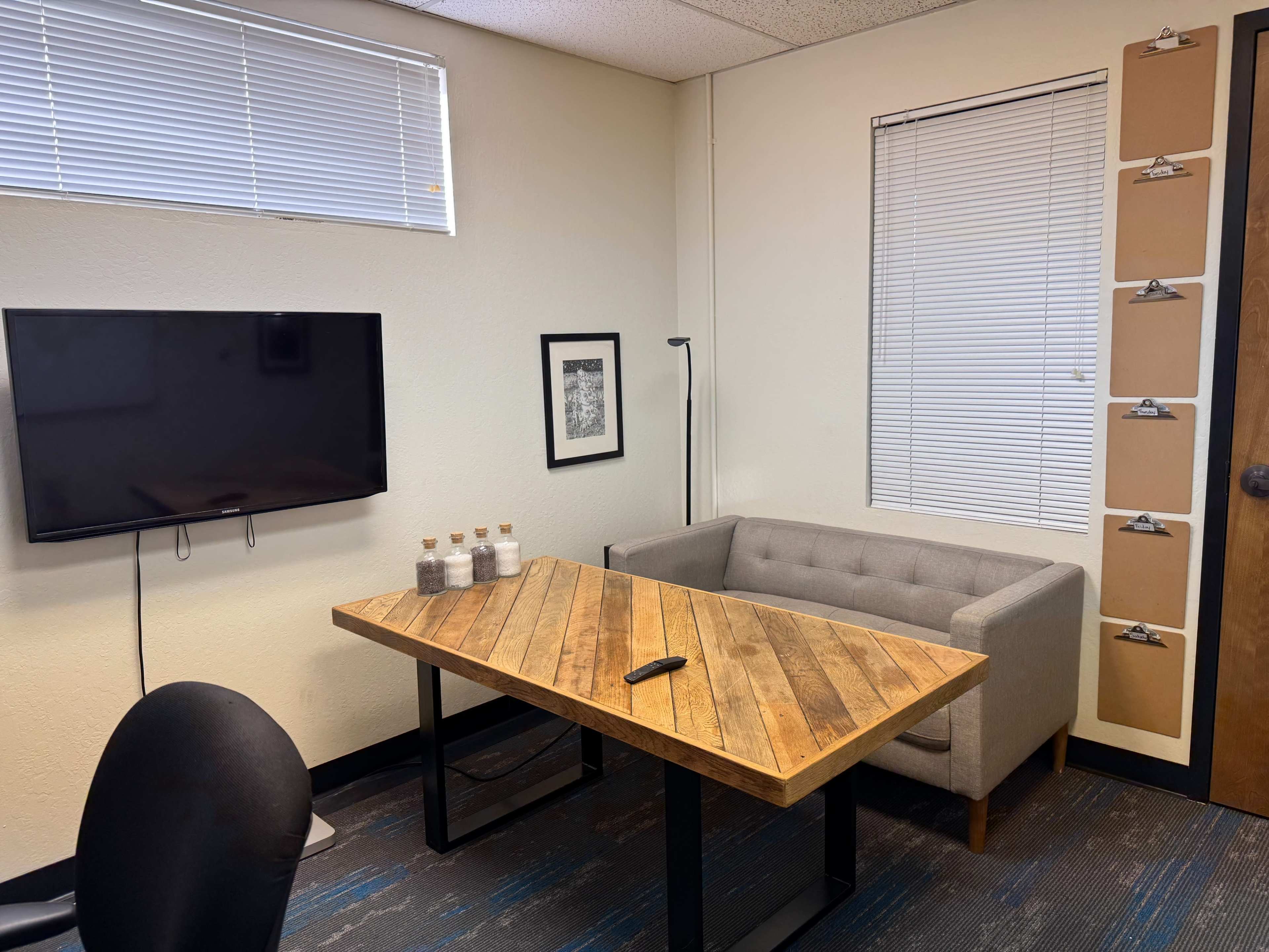 The image shows a small office with a wooden table, a gray couch, a wall-mounted TV, and several clipboards on the wall.