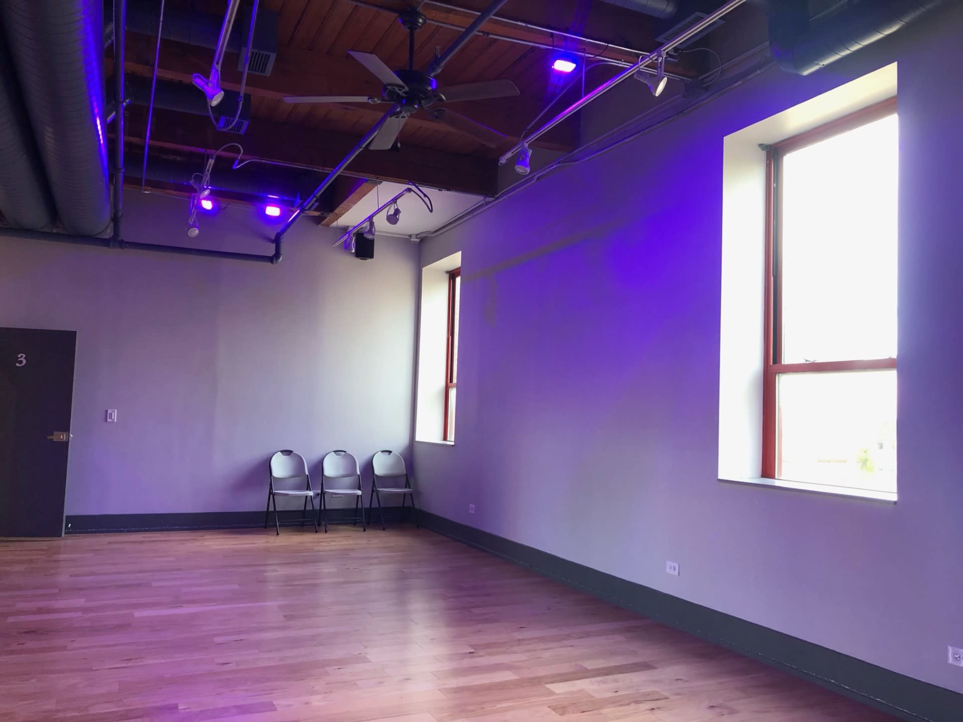 An empty room with wooden flooring, two large windows, and a row of four folding chairs against a wall.