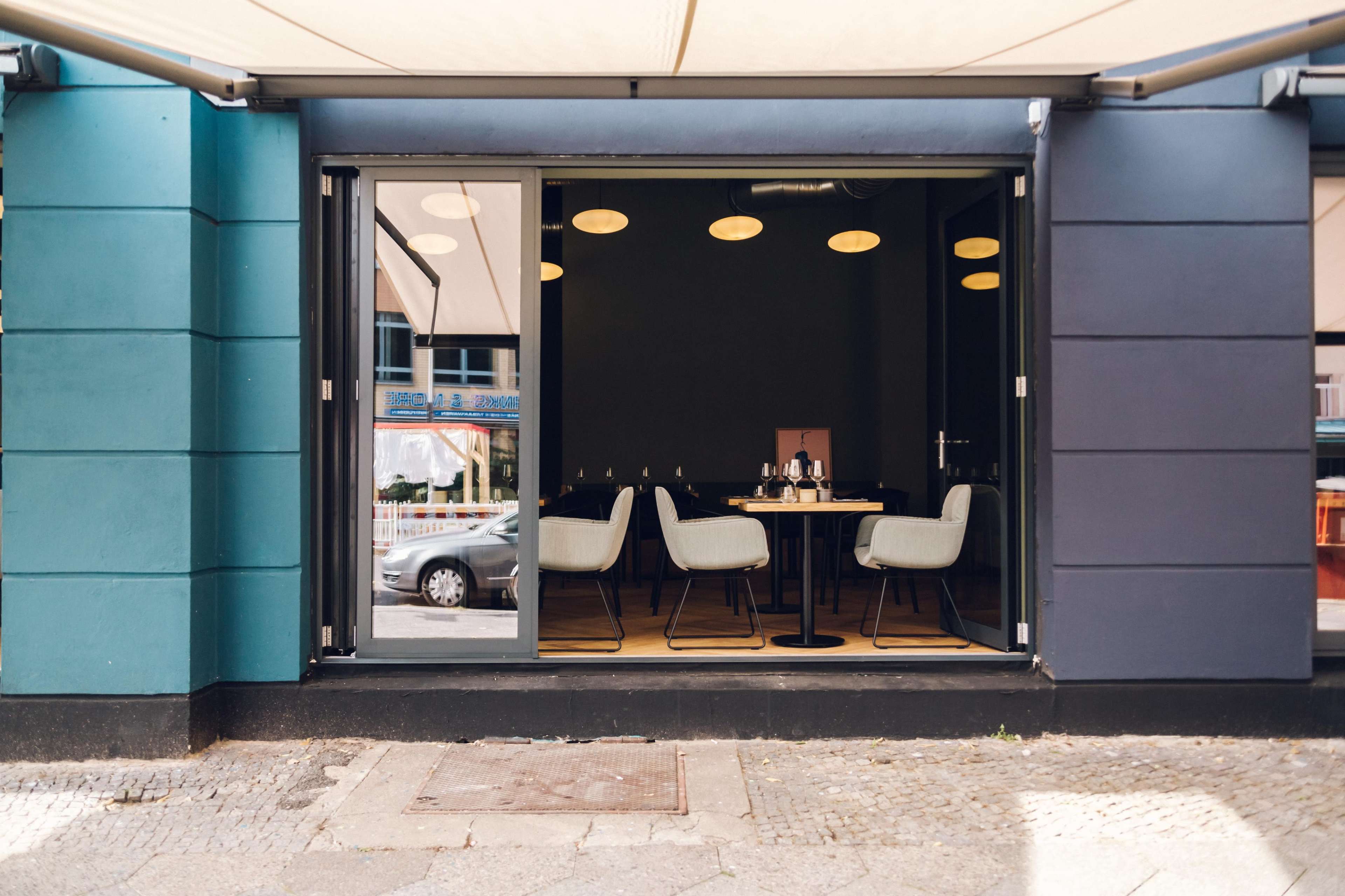 The image shows a dining area with modern chairs and tables visible through an open window in a building with blue and black exterior walls.