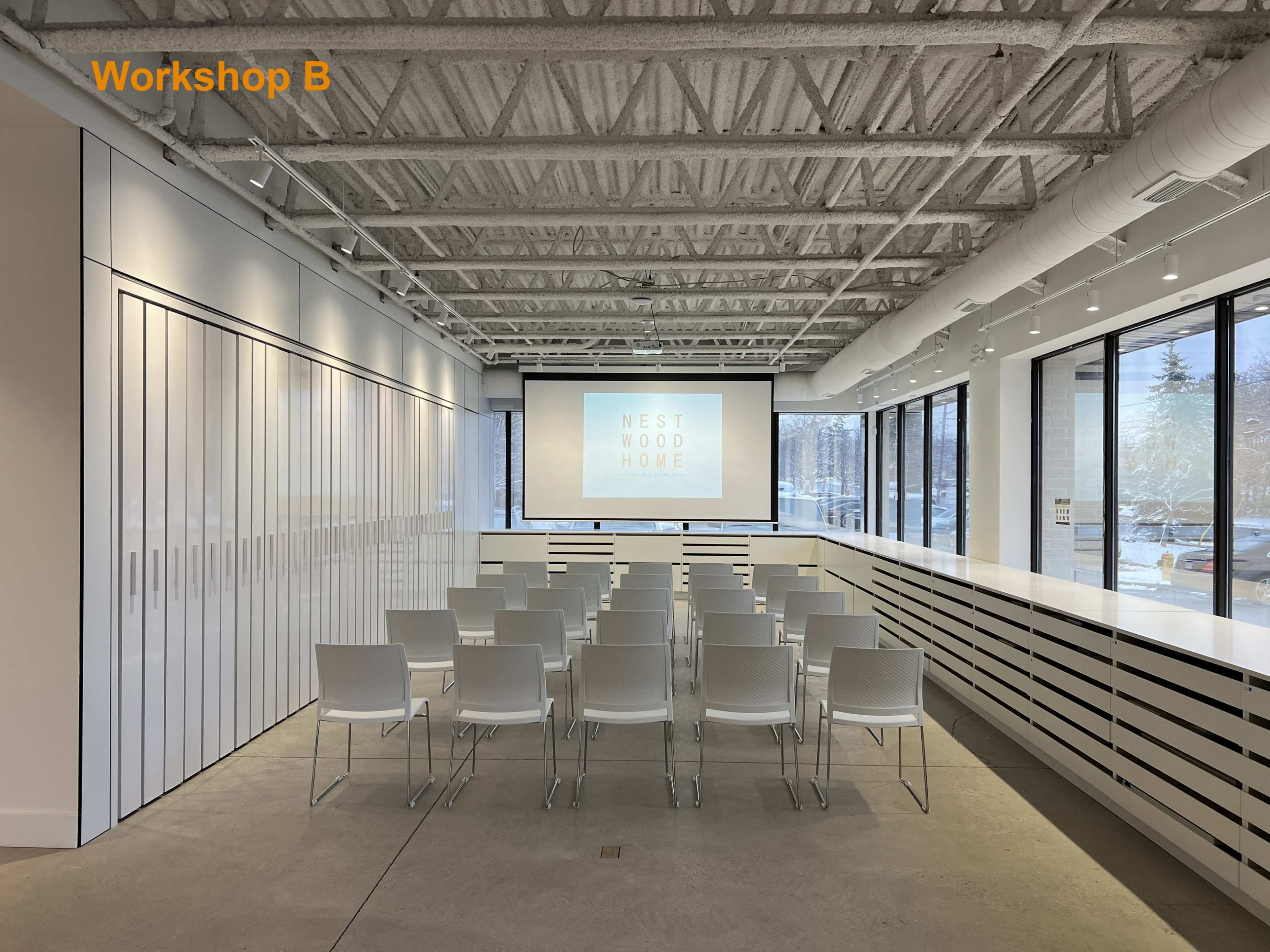 A modern workshop space with rows of white chairs facing a large screen displaying the text "NEST WOOD HOME."