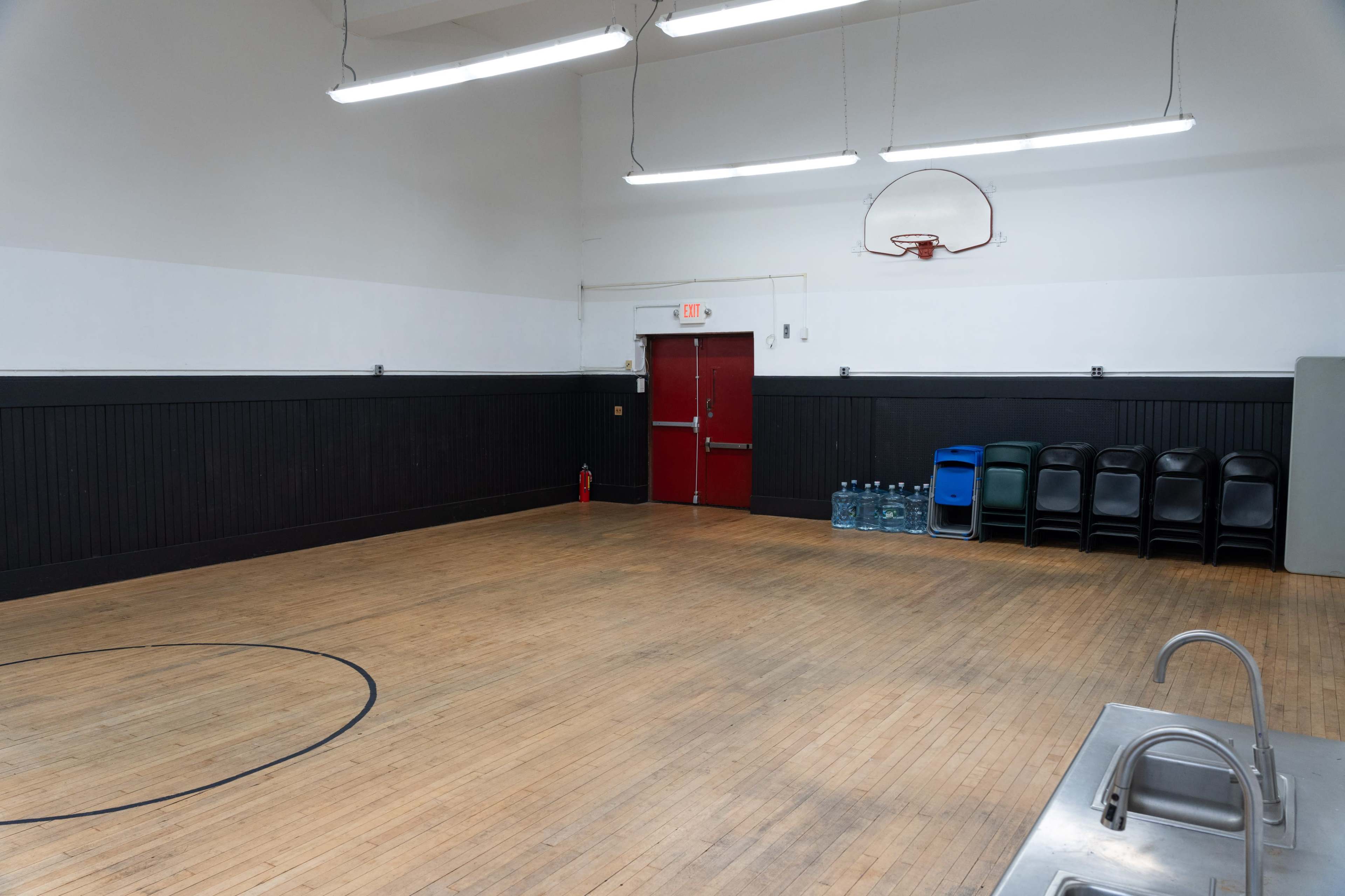The image shows an empty gymnasium with a basketball hoop on the wall, wooden flooring, and a row of black chairs and water containers against the back wall.