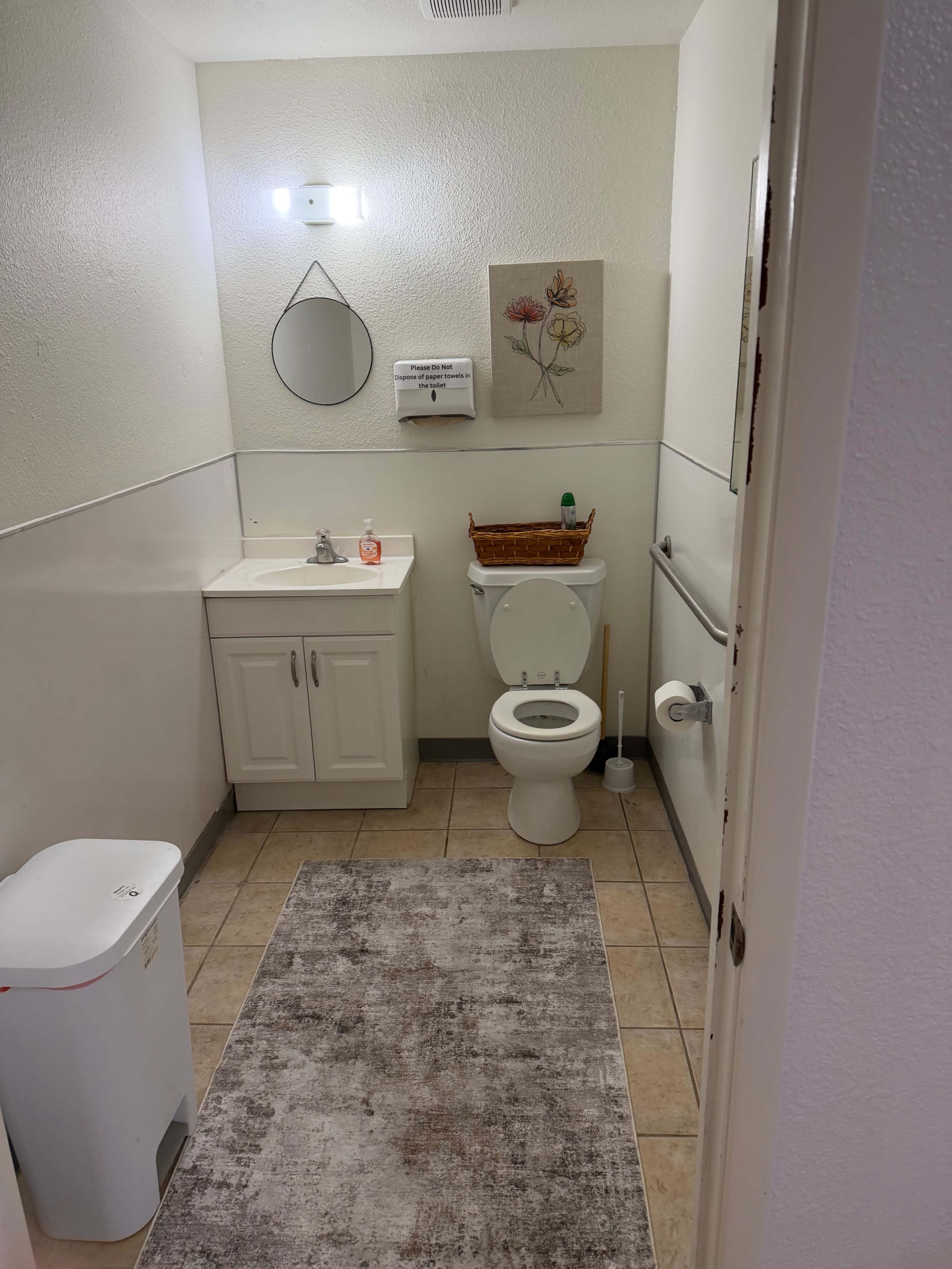 The image shows a small bathroom with a toilet, a sink with a cabinet underneath, a wall mirror, and light-colored tiles on the floor.