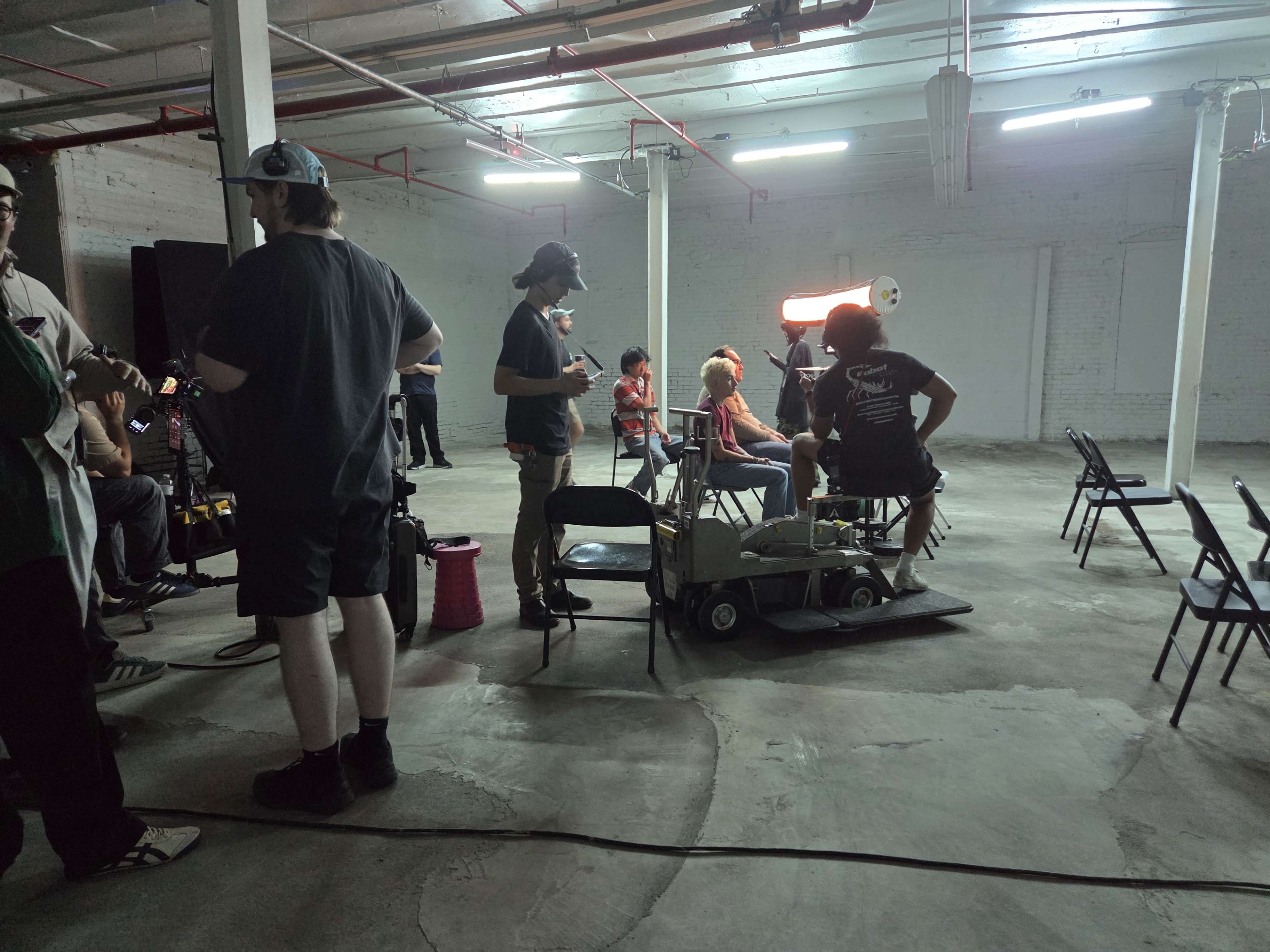 A film crew sets up equipment and prepares for a shoot in a spacious, empty industrial area with concrete floors and exposed overhead pipes.