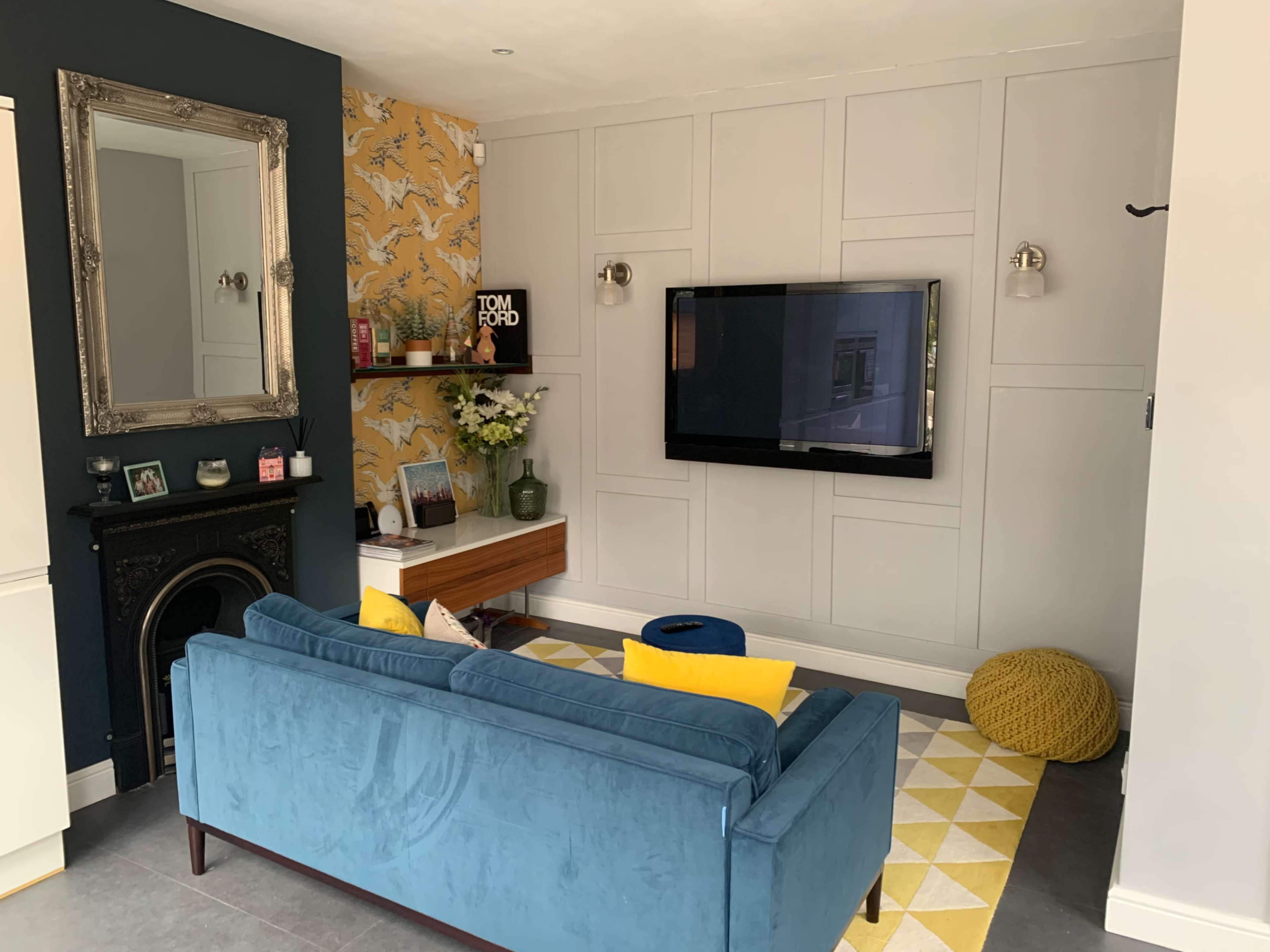 The living room features a blue velvet sofa, a mounted television on a light gray wall, and a patterned yellow and white rug.