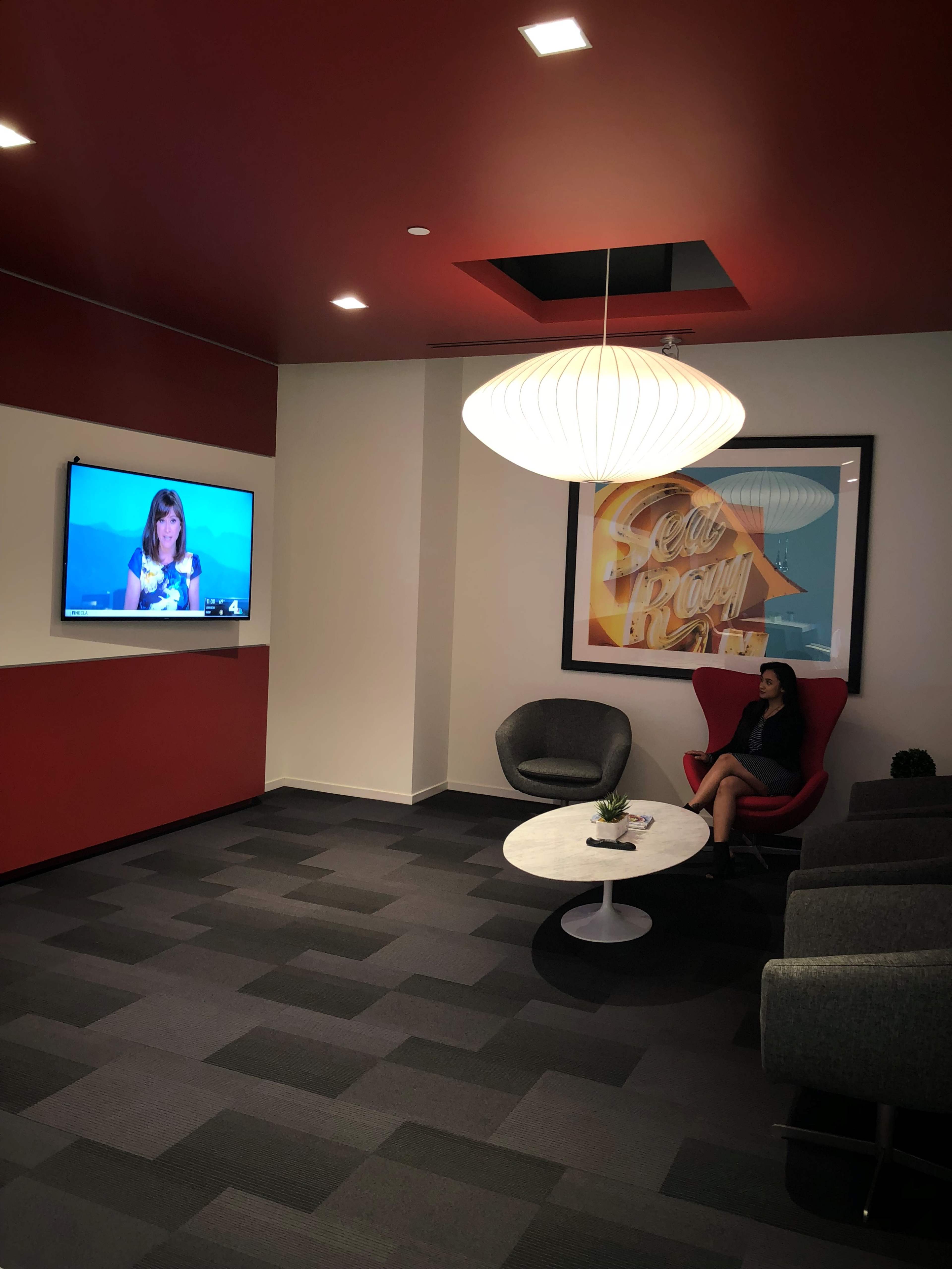 A woman sits in a modern lounge area with a television displaying a video call on one wall and a large framed photo on another wall.