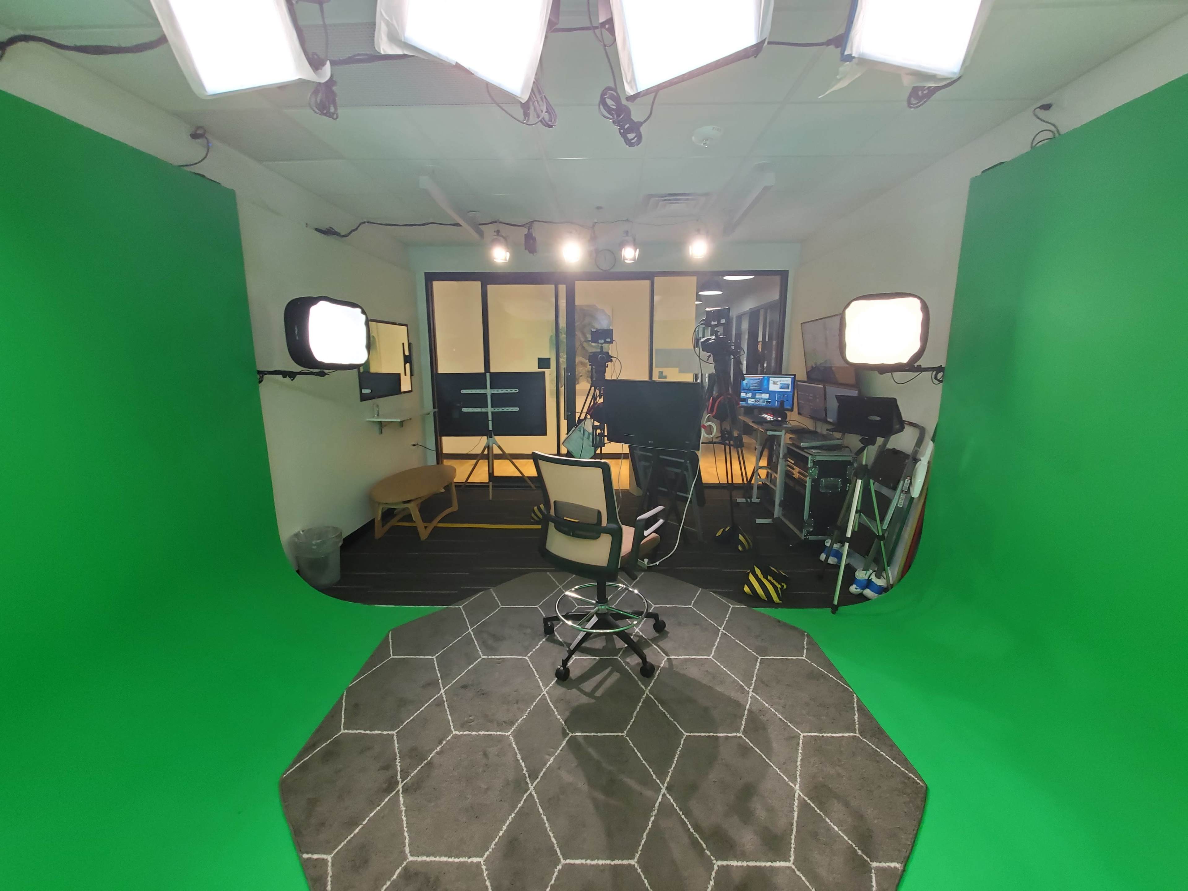 The image shows a video recording studio with a green screen backdrop, bright lights, and a central chair on a hexagonal-patterned floor.