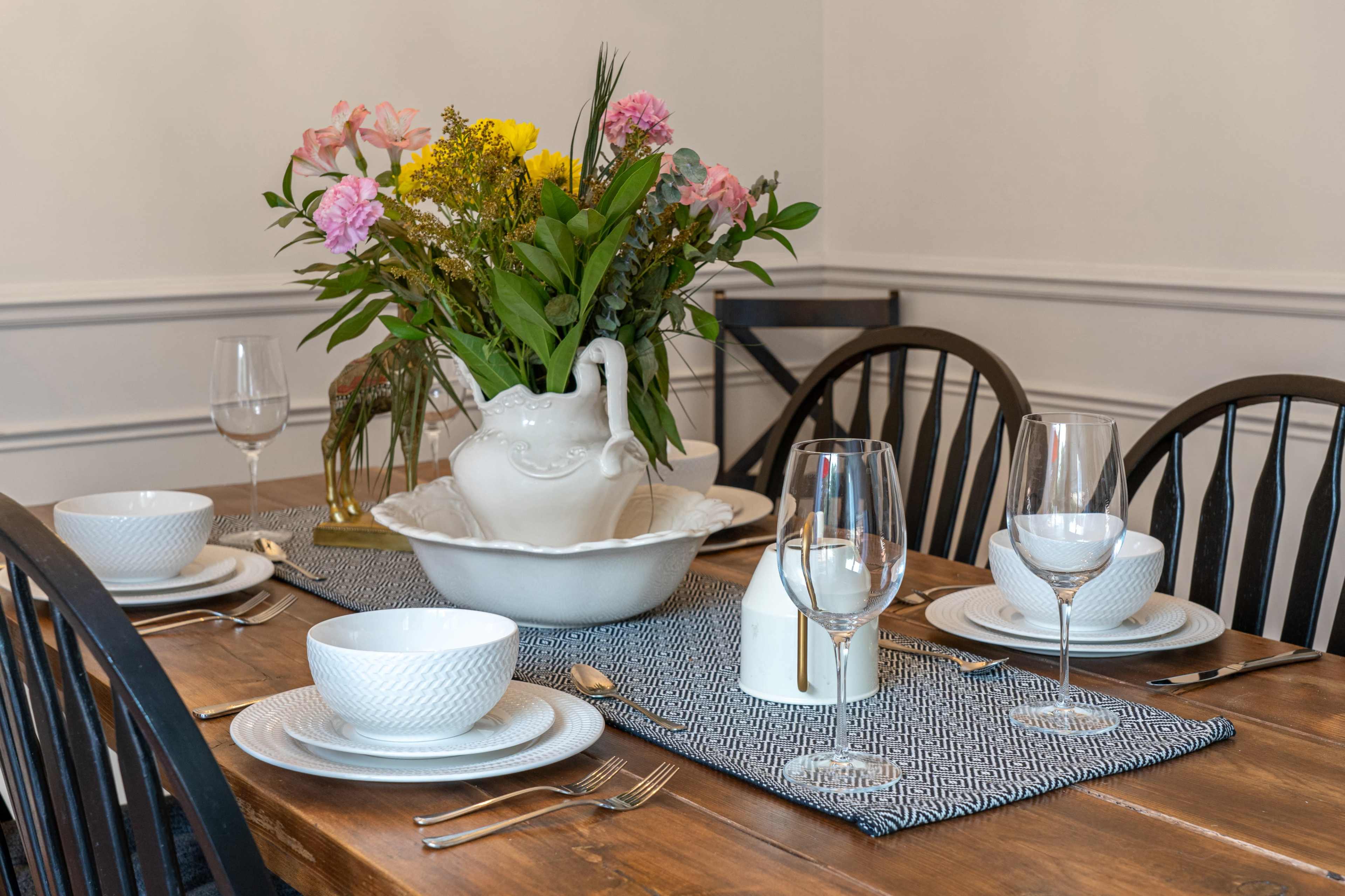 A dining table is set with white dishware and glassware, featuring a centerpiece of colorful flowers in a decorative vase.