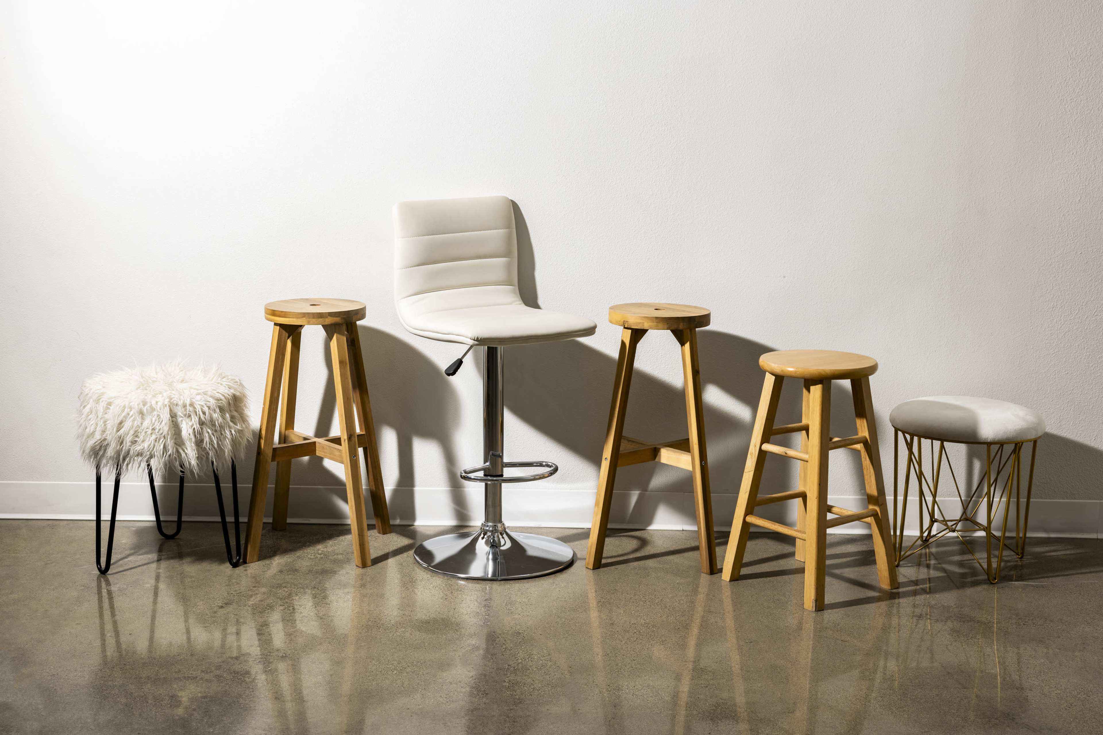 The image features a variety of five stools arranged against a plain wall, including three wooden stools, one white bar stool, and one stool with a fluffy seat.