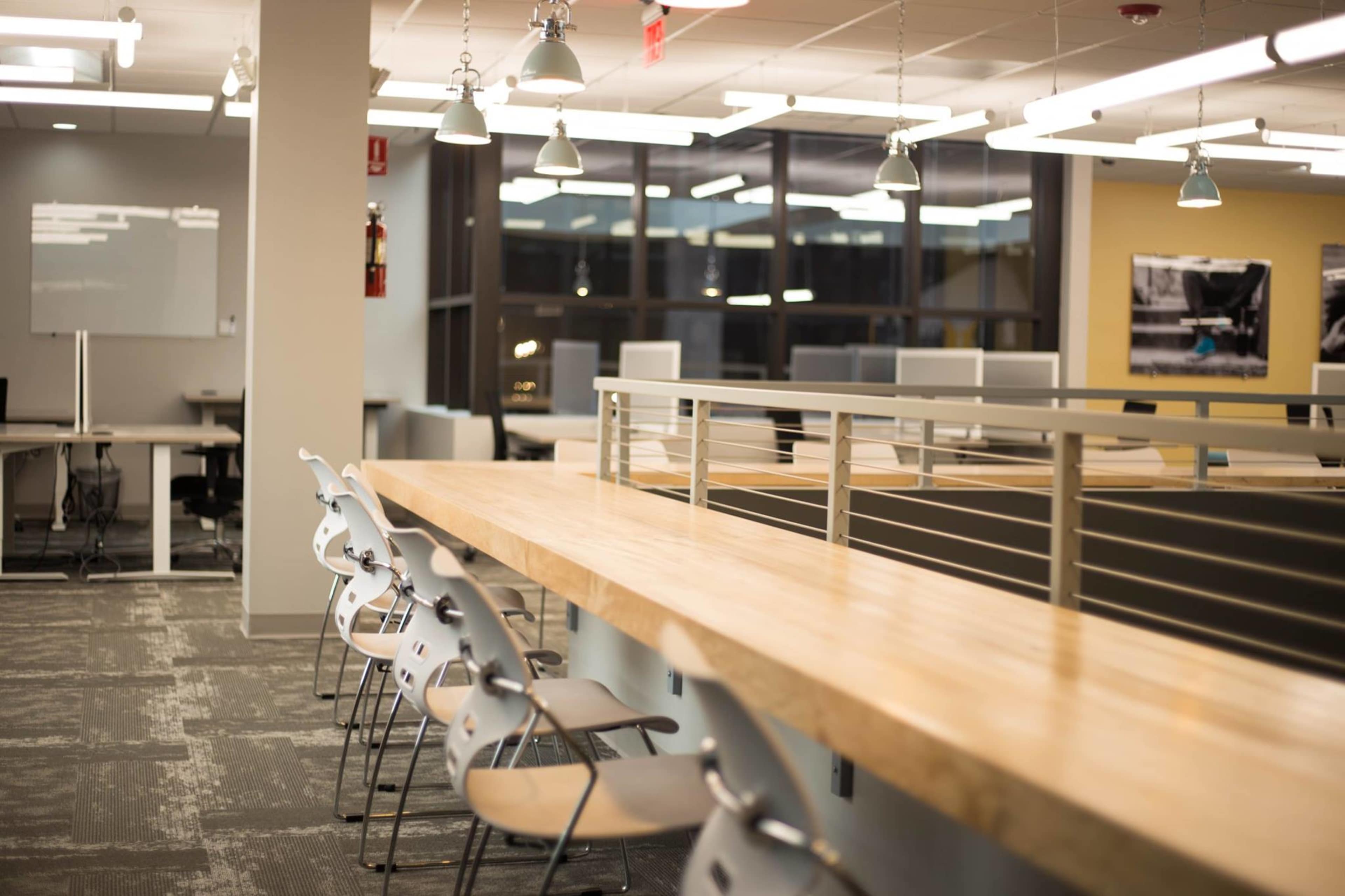 A modern office space with a long wooden countertop and several empty gray chairs in a well-lit environment.