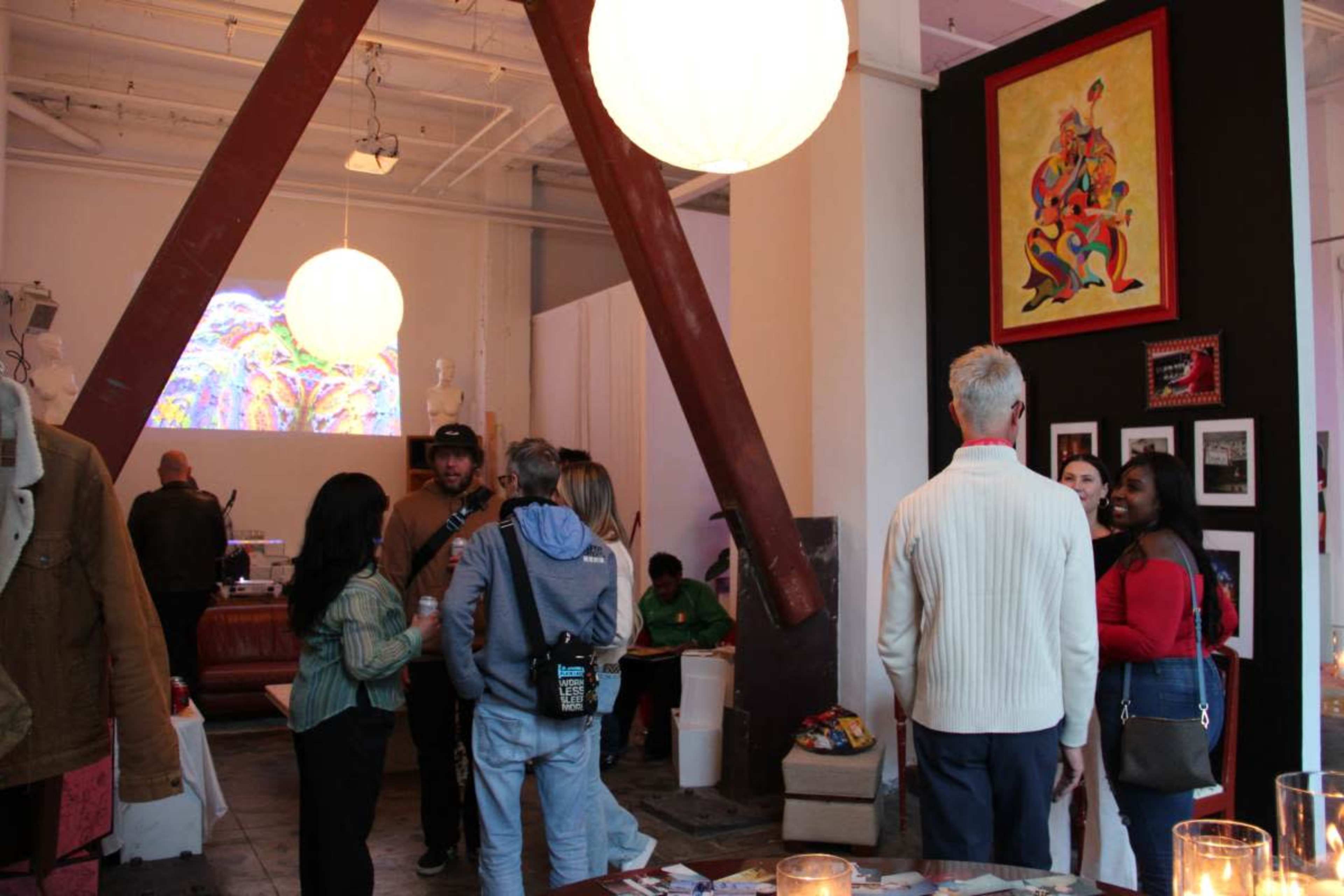 The image shows a spacious art gallery with people socializing, viewing artwork on the walls, and large hanging lights overhead.