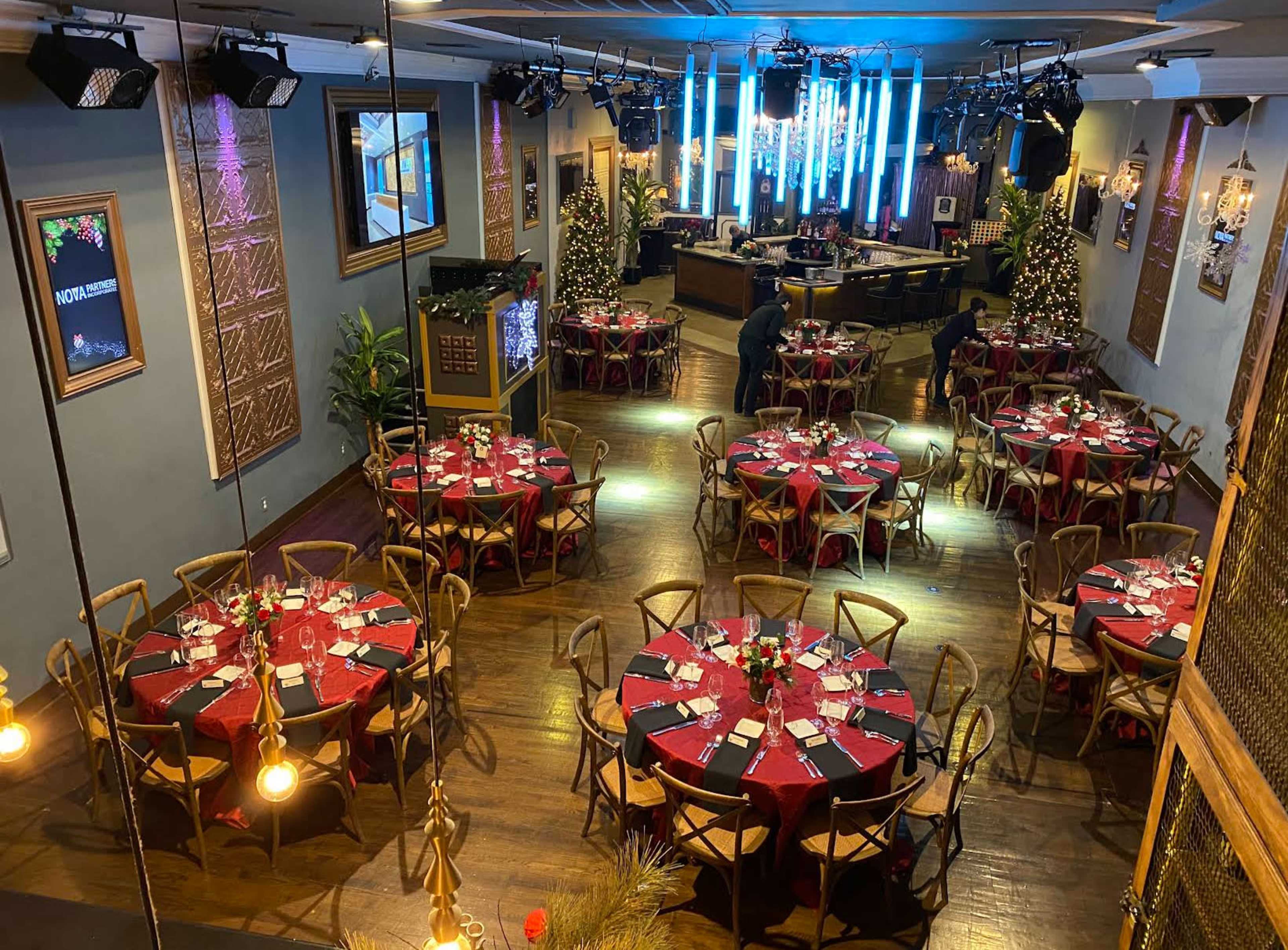 The image shows a spacious dining area with rows of round tables set with red tablecloths and neatly arranged plates and cutlery, illuminated by overhead lights and decorated for an event.