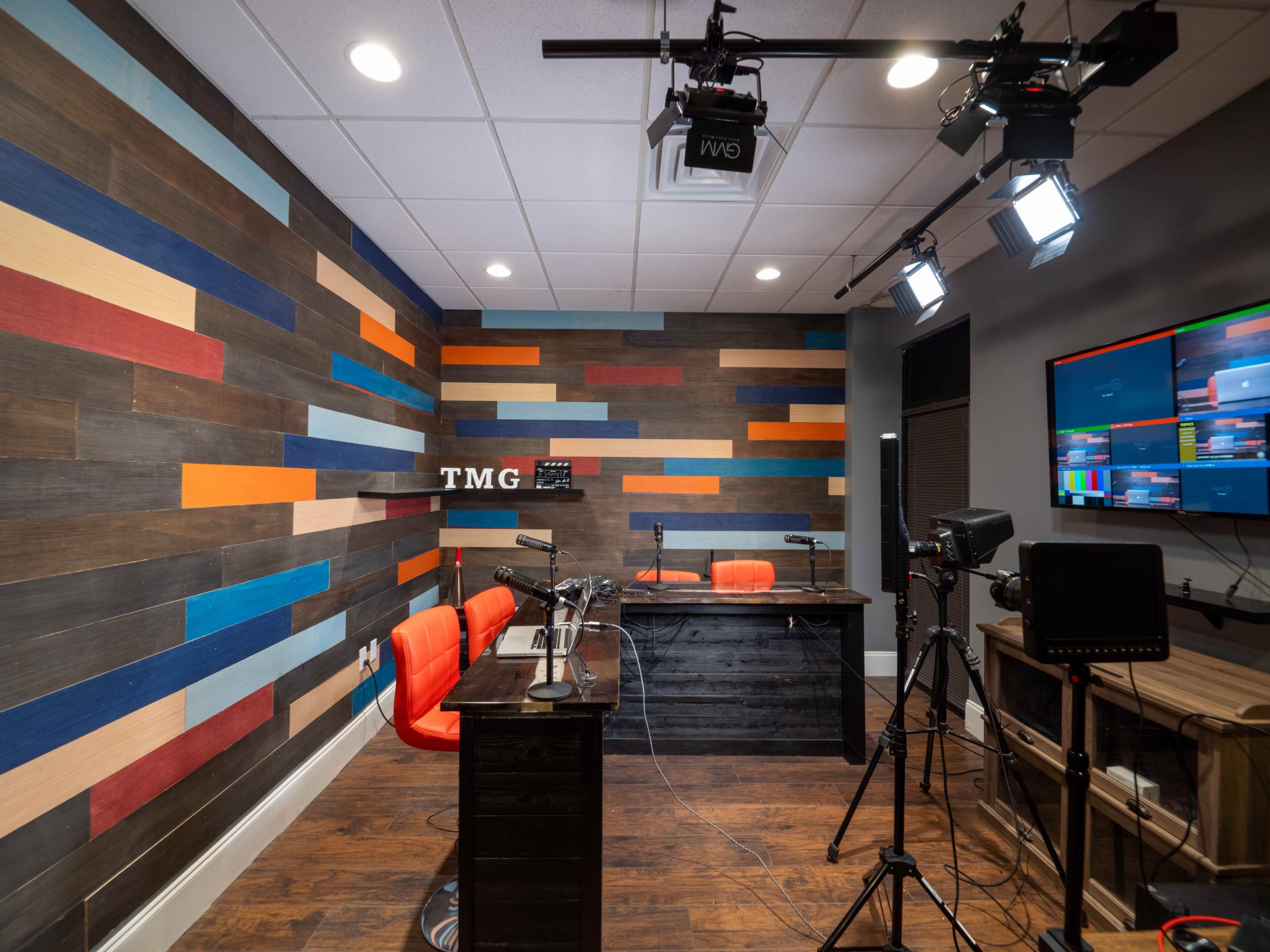 A modern studio setup with a wooden desk, two orange chairs, and colorful wooden paneling on the walls, complete with lighting equipment and a monitor displaying video feeds.