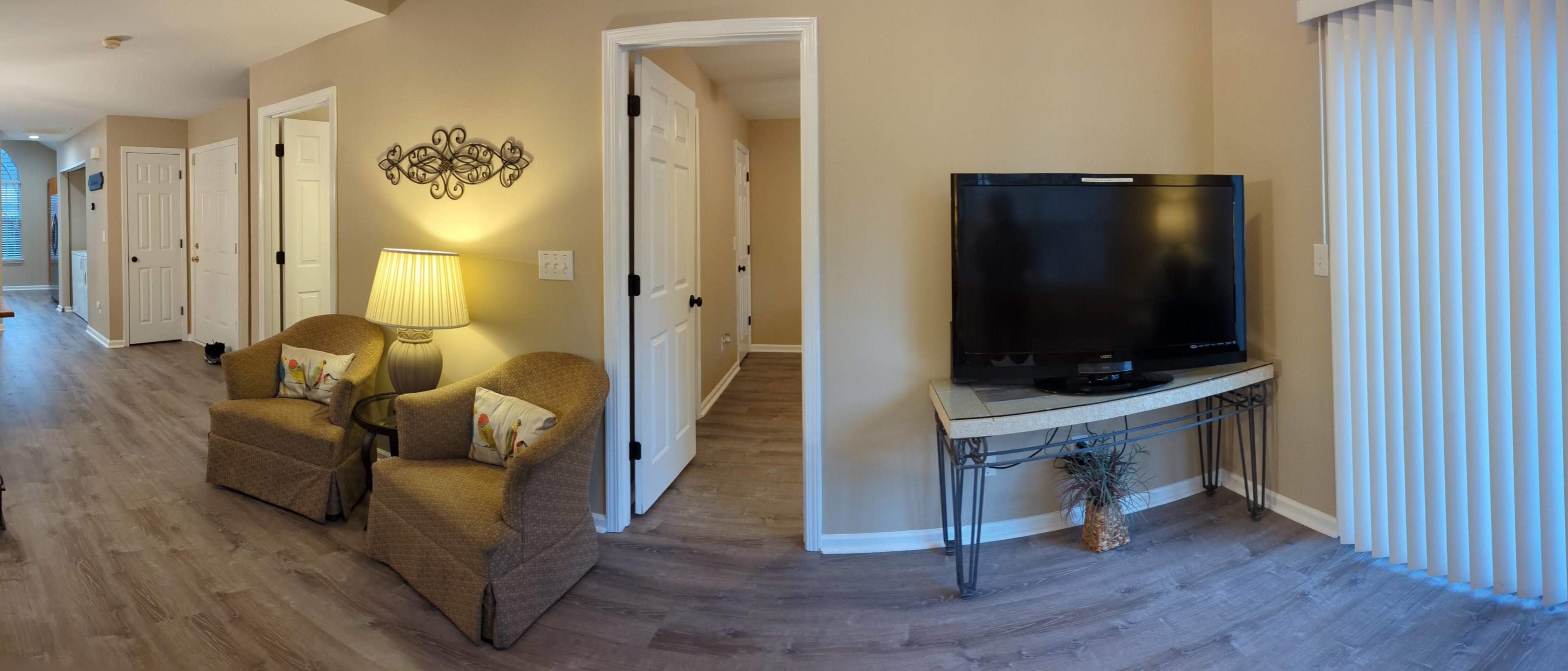 The image shows a cozy living area featuring two patterned chairs, a lamp, a television on a small table, and light-colored walls.
