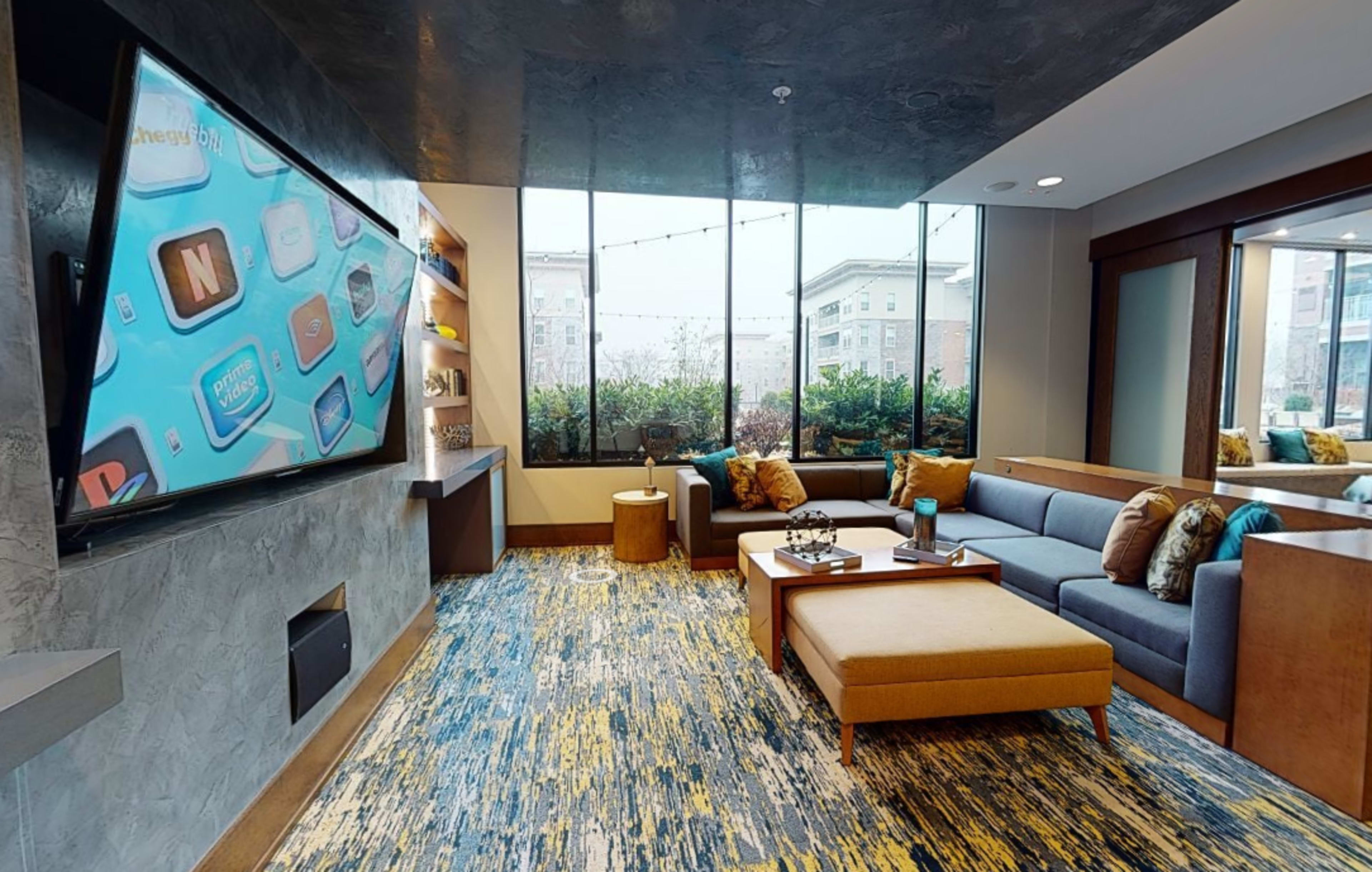 A modern lounge area featuring a large television display, a sectional sofa, and a coffee table, surrounded by large windows with a view of the outdoors.