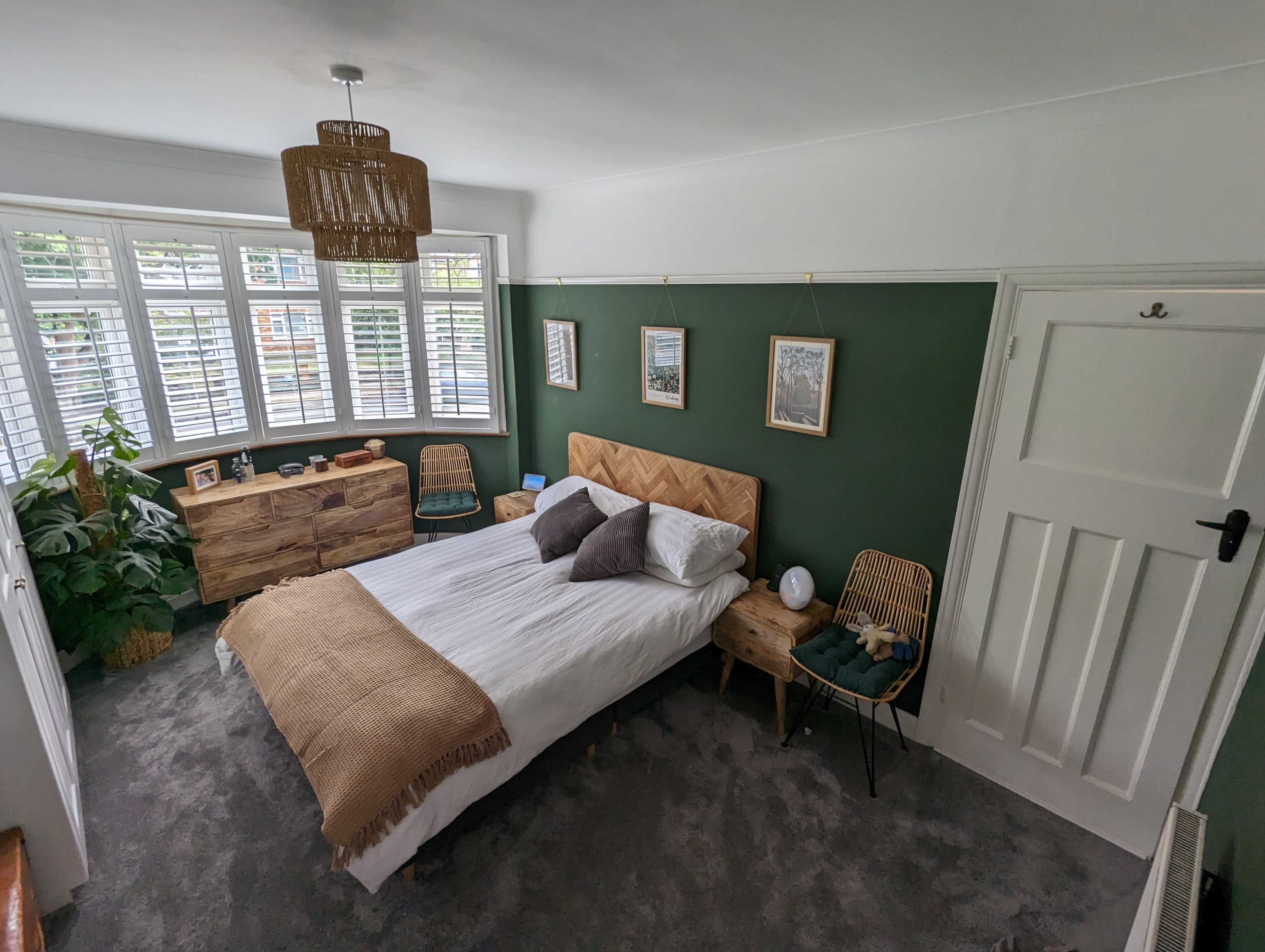A neatly arranged bedroom features a green accent wall, a wooden bed frame, and a cozy seating area by a bay window.