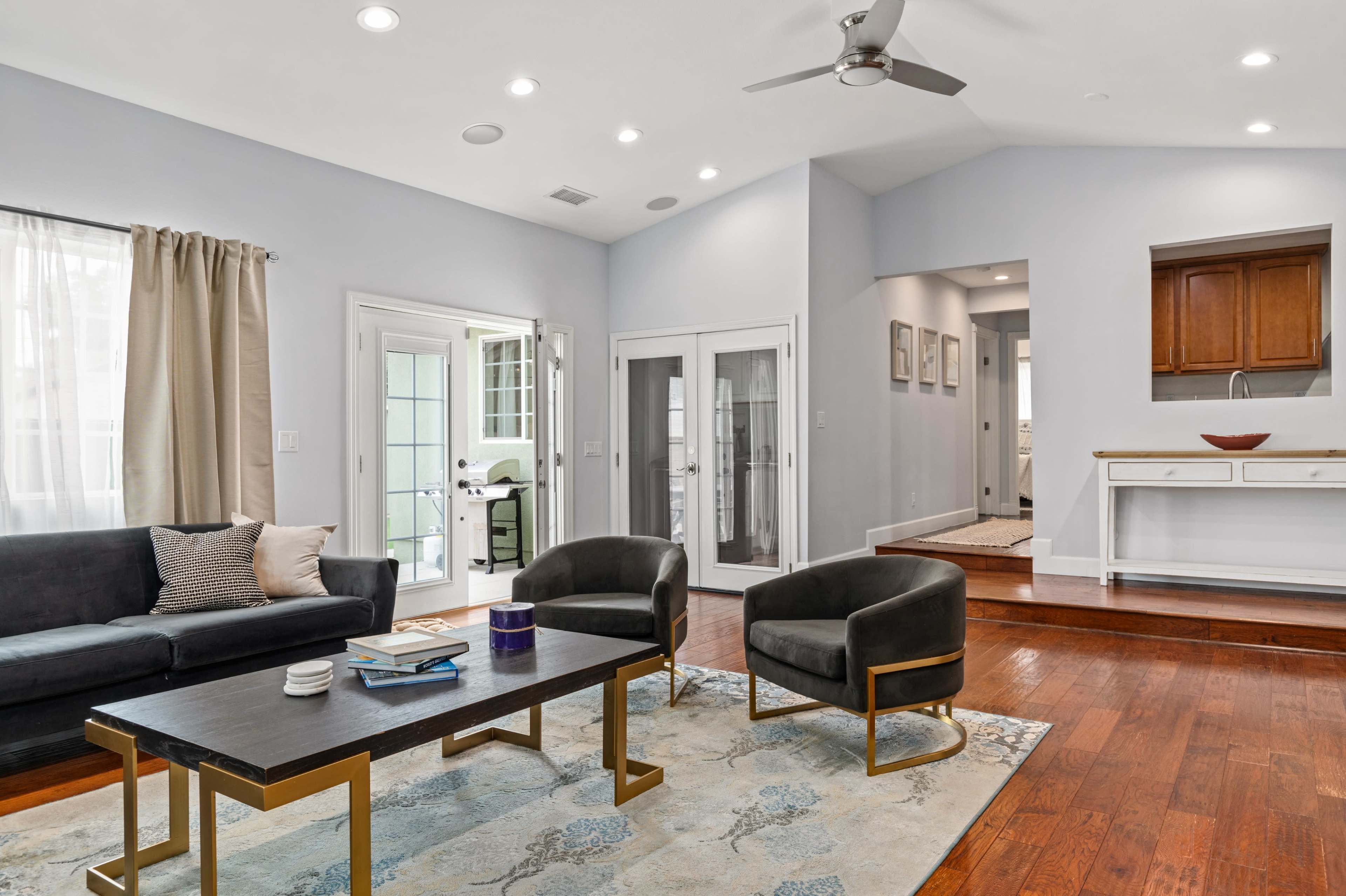 A modern living room with a dark sofa, two chairs, a rectangular coffee table, and a doorway leading to a bright area with a view of a kitchen.