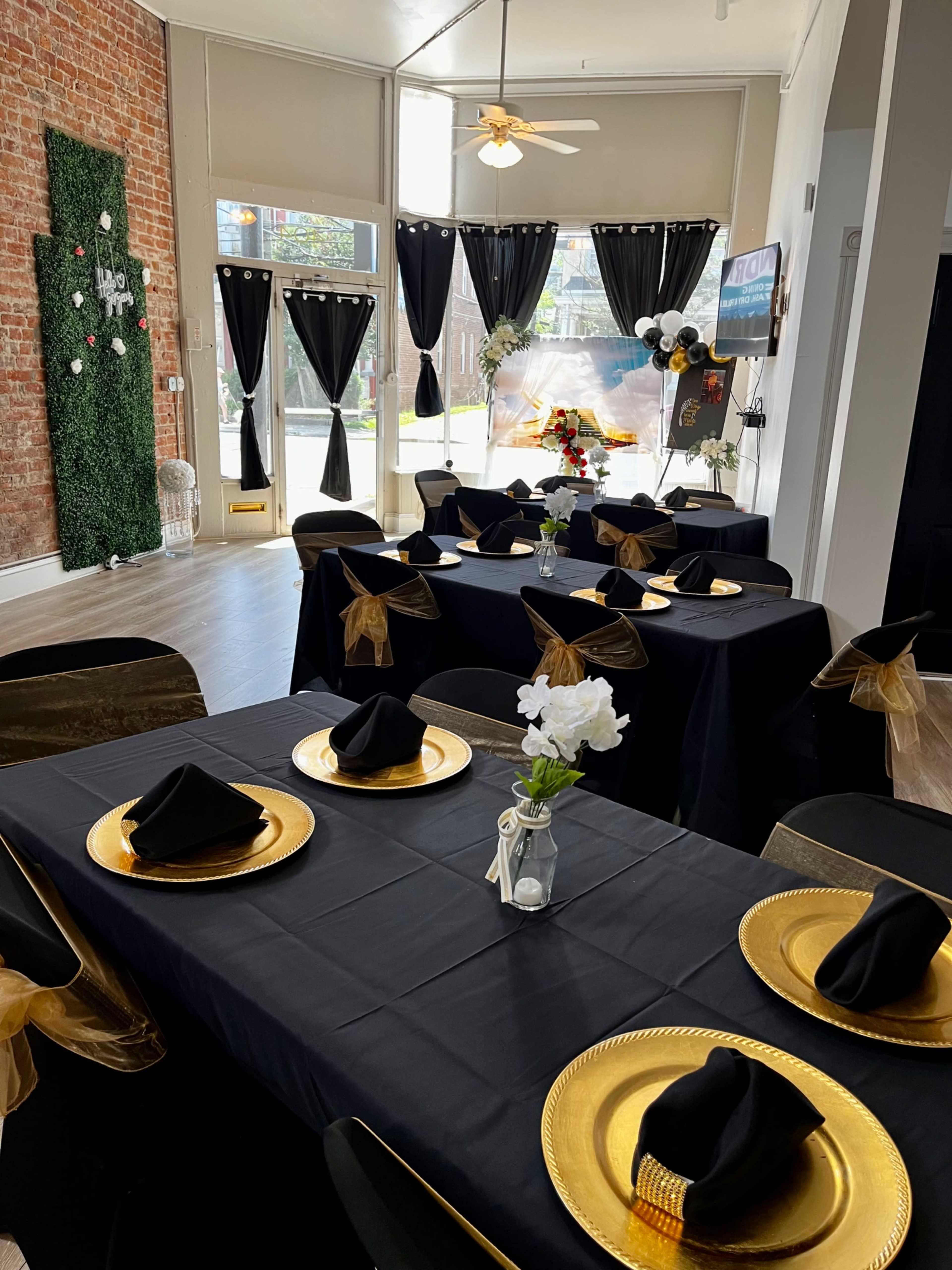 A dining area is set up with black tablecloths, gold chargers, and neatly folded black napkins at several tables, with large windows draped in black curtains in the background.