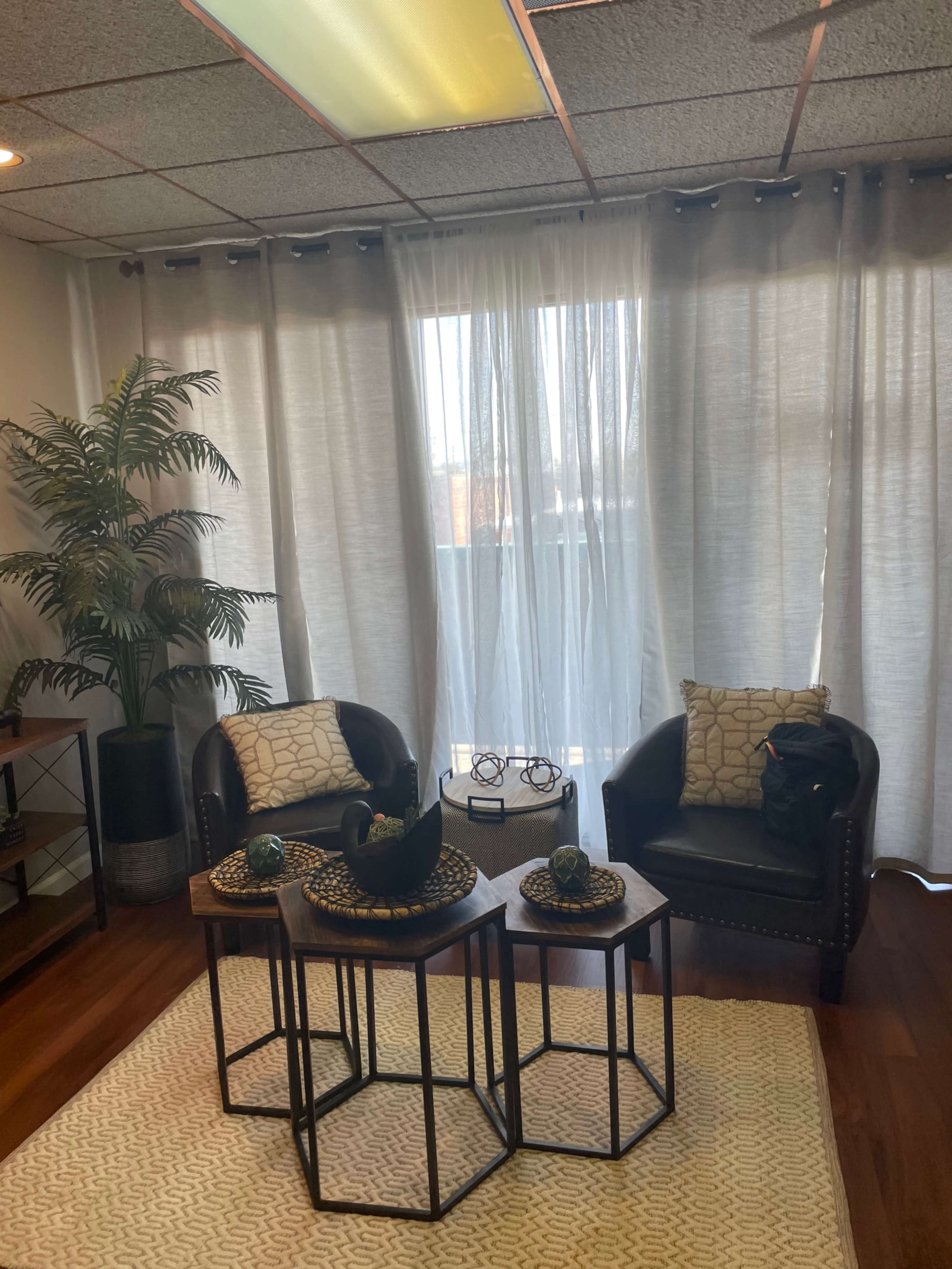 A cozy room with two black chairs, a set of three nested tables, a plant in the corner, and sheer curtains letting in light.