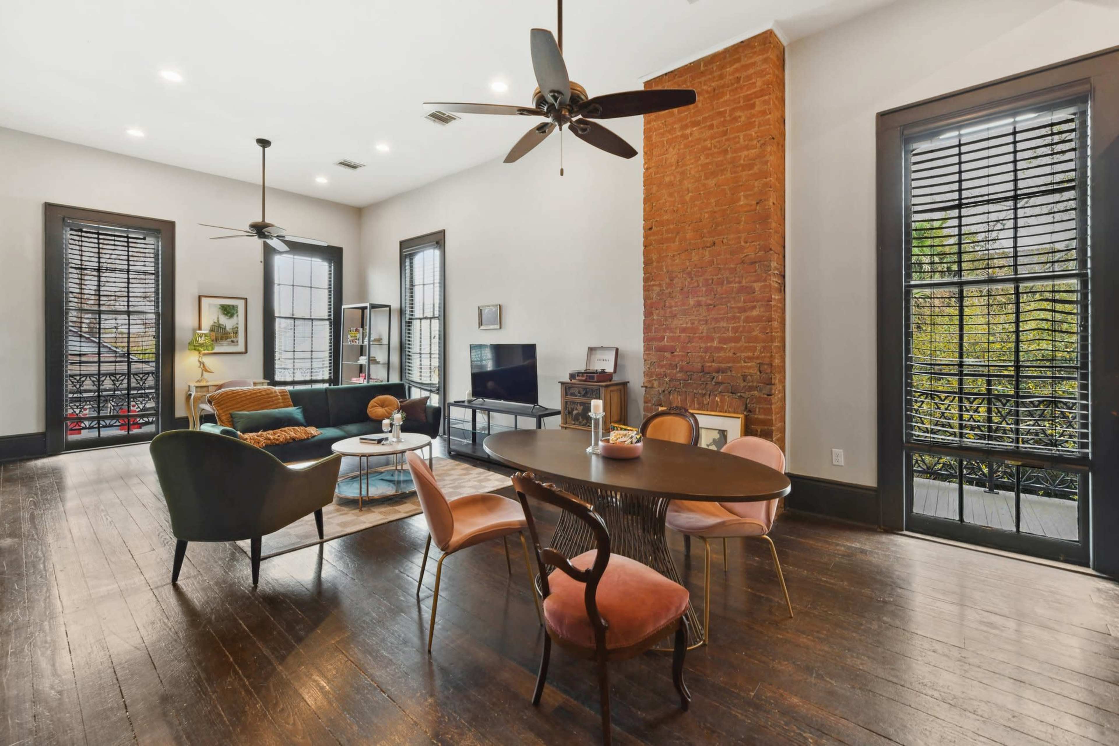 A spacious room features a dining table, chairs, a couch, and a television, with exposed brick walls and large windows adorned with shutters.
