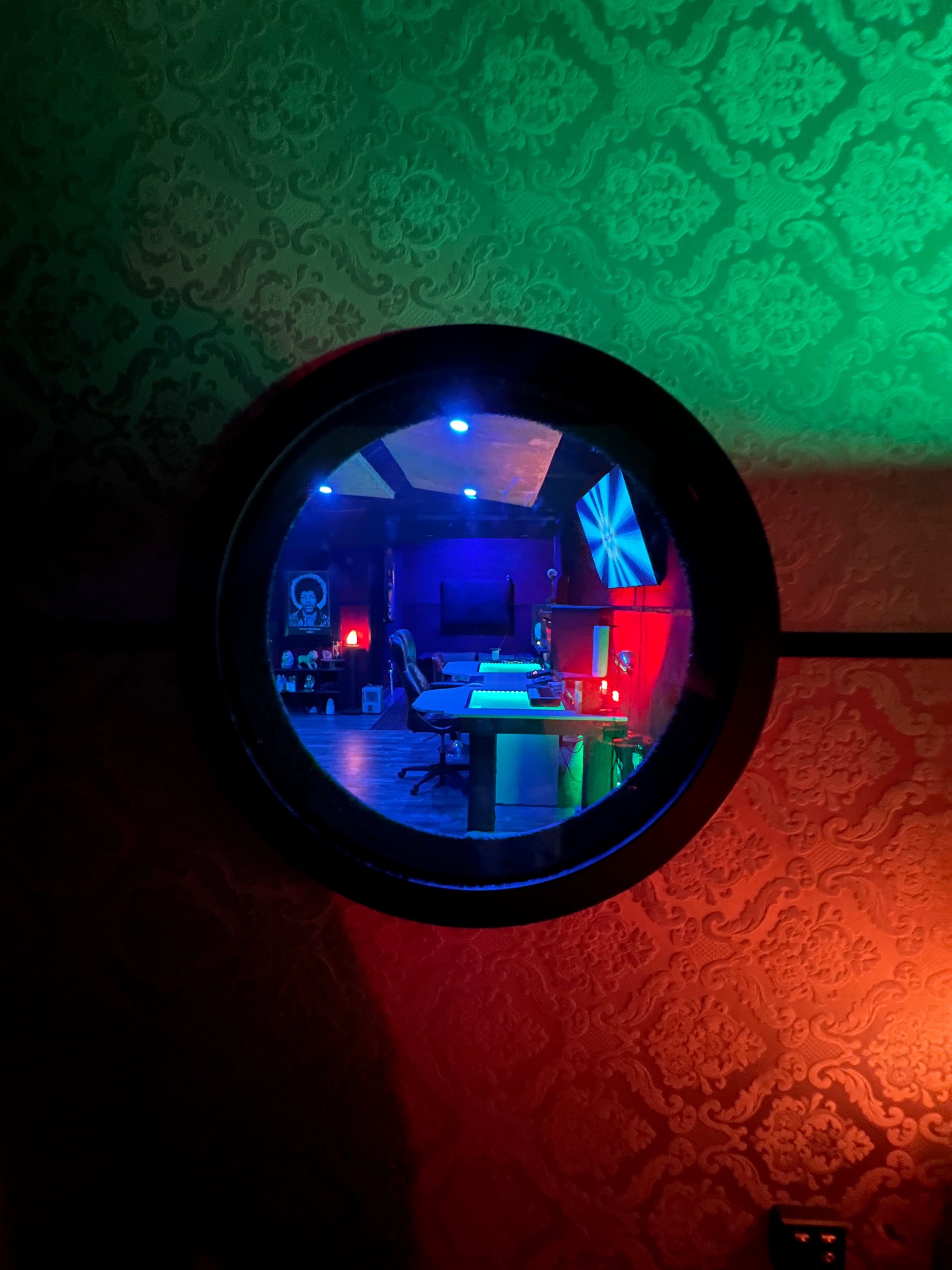 A circular window view into a dimly lit recording studio with colorful lighting and various music equipment.