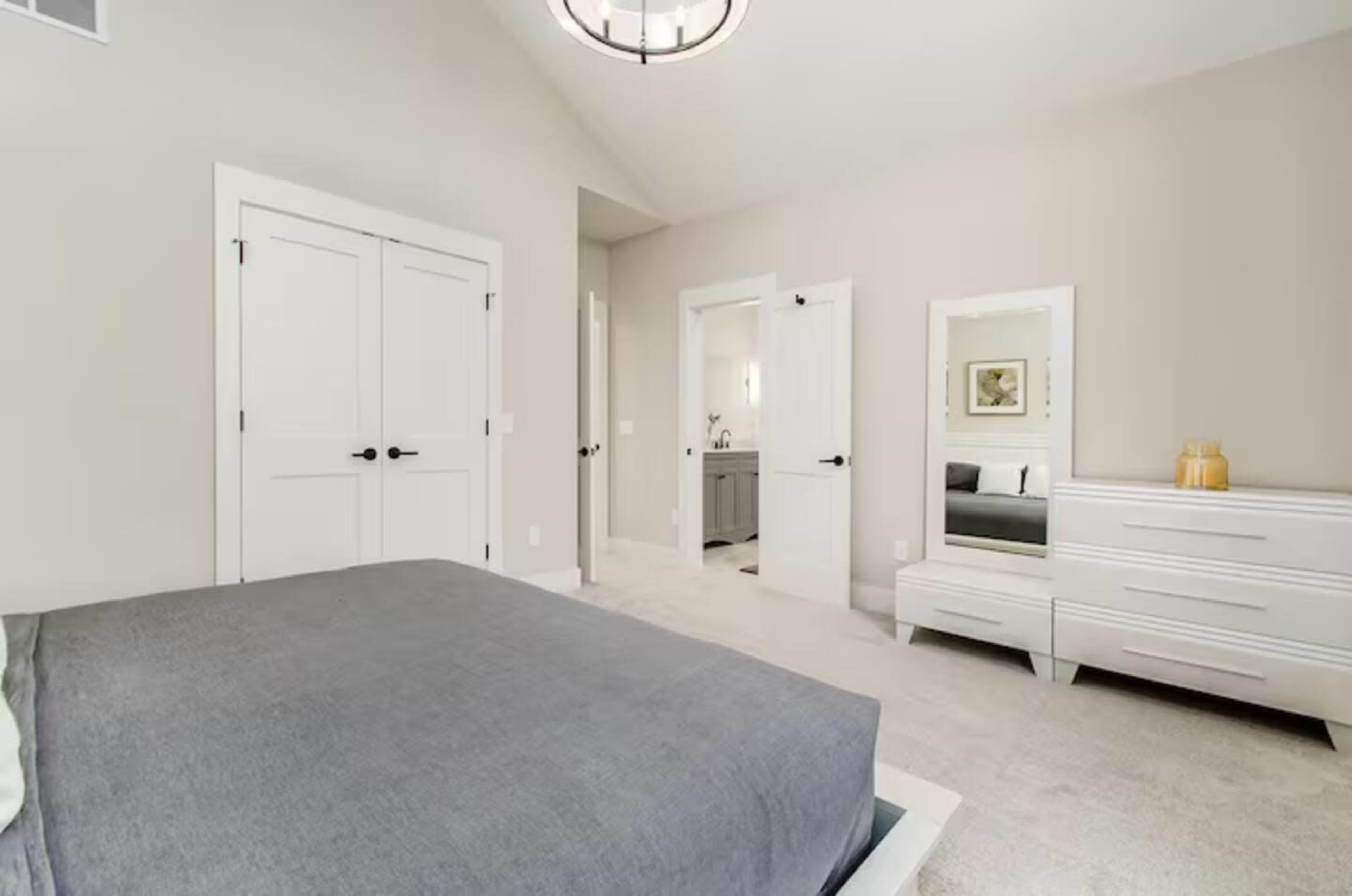 The image shows a spacious, modern bedroom featuring a large bed, a dresser, a mirror, and double doors leading to a bathroom.