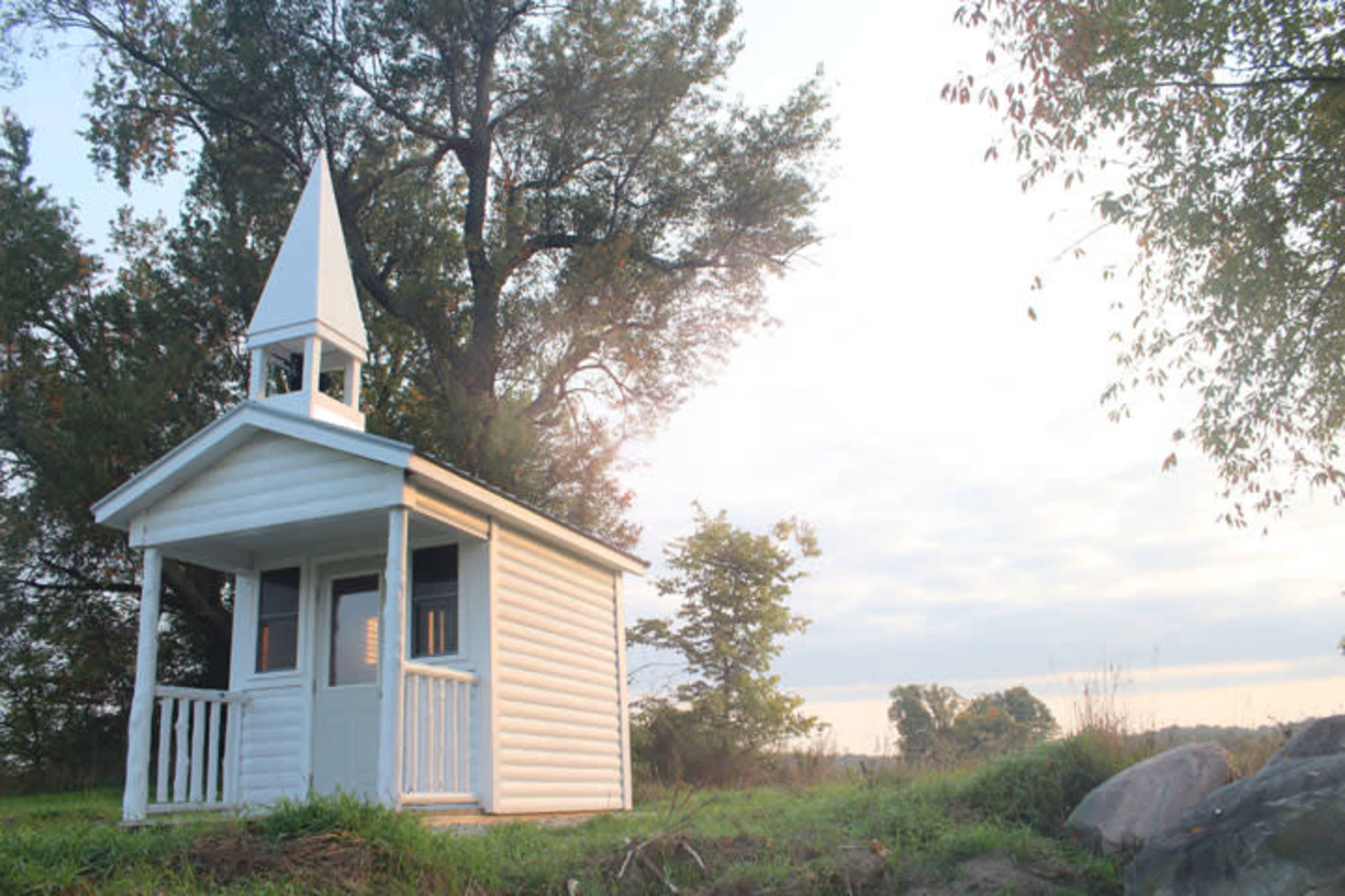 A small white chapel with a spire stands next to a tree under a cloudy sky.