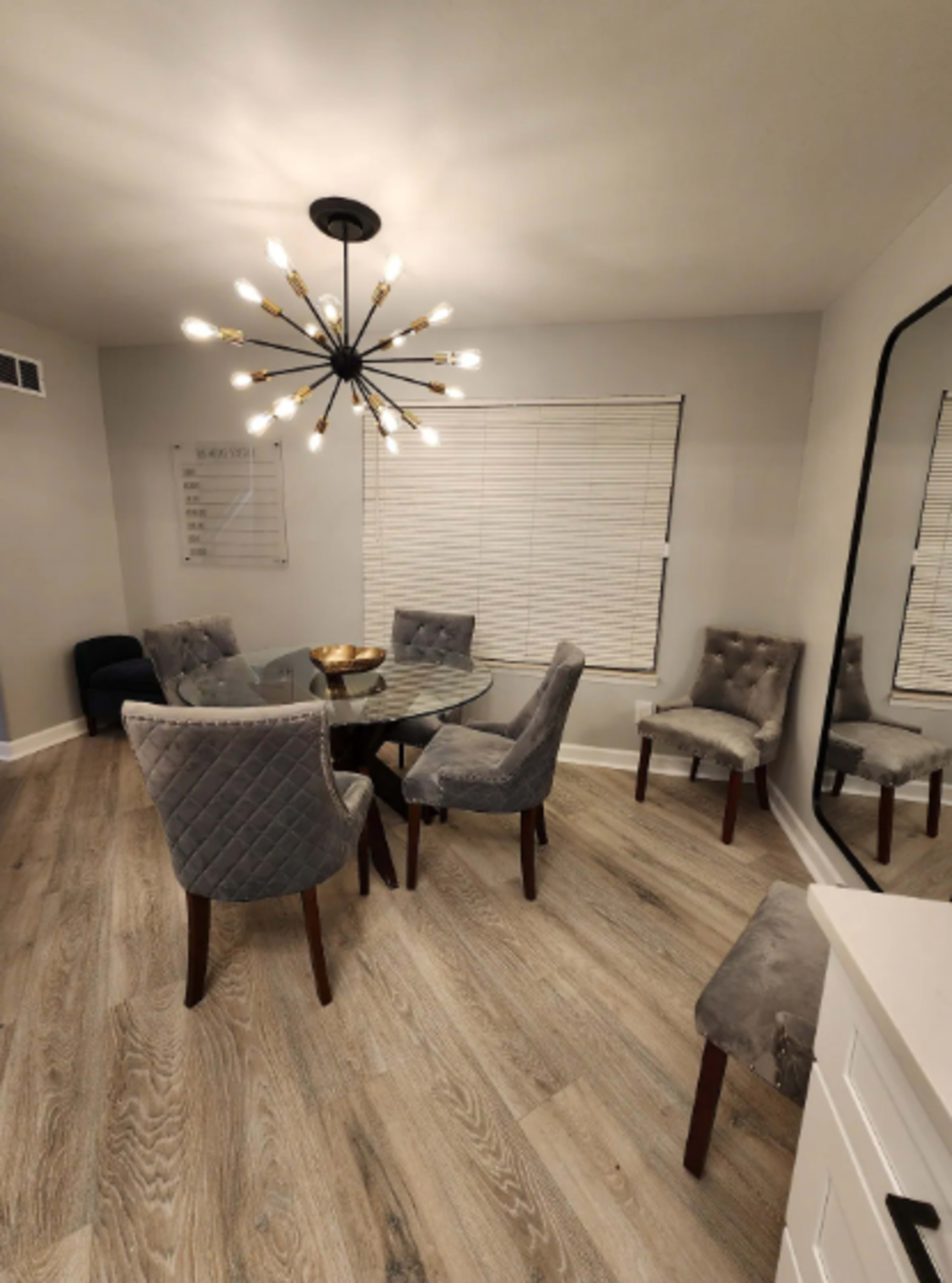A modern dining area features a round glass table surrounded by six upholstered chairs, with a large chandelier and a mirror on the wall.