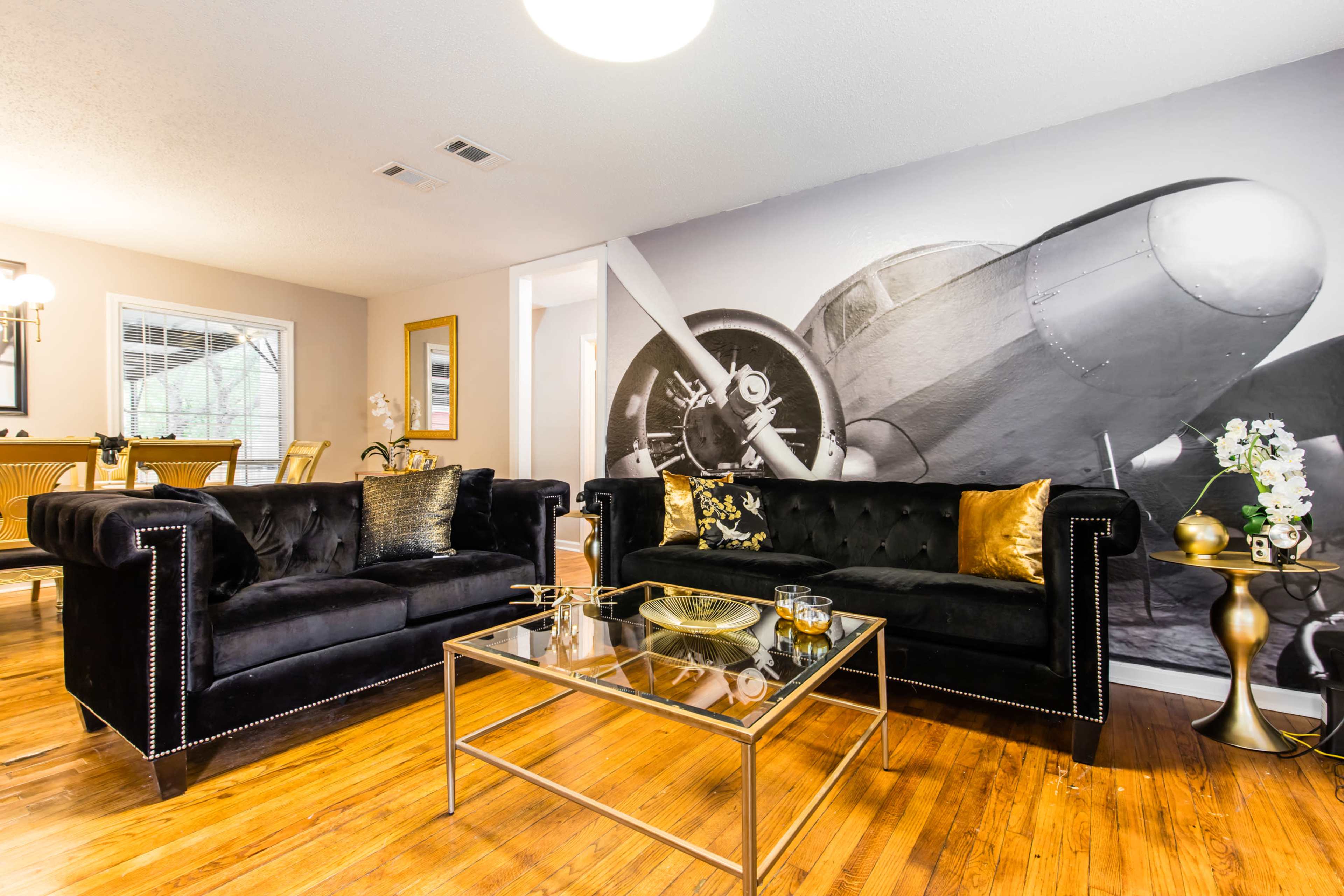 The scene features a living room with two black velvet sofas positioned in front of a large mural of an airplane, accompanied by a glass coffee table and gold accents.